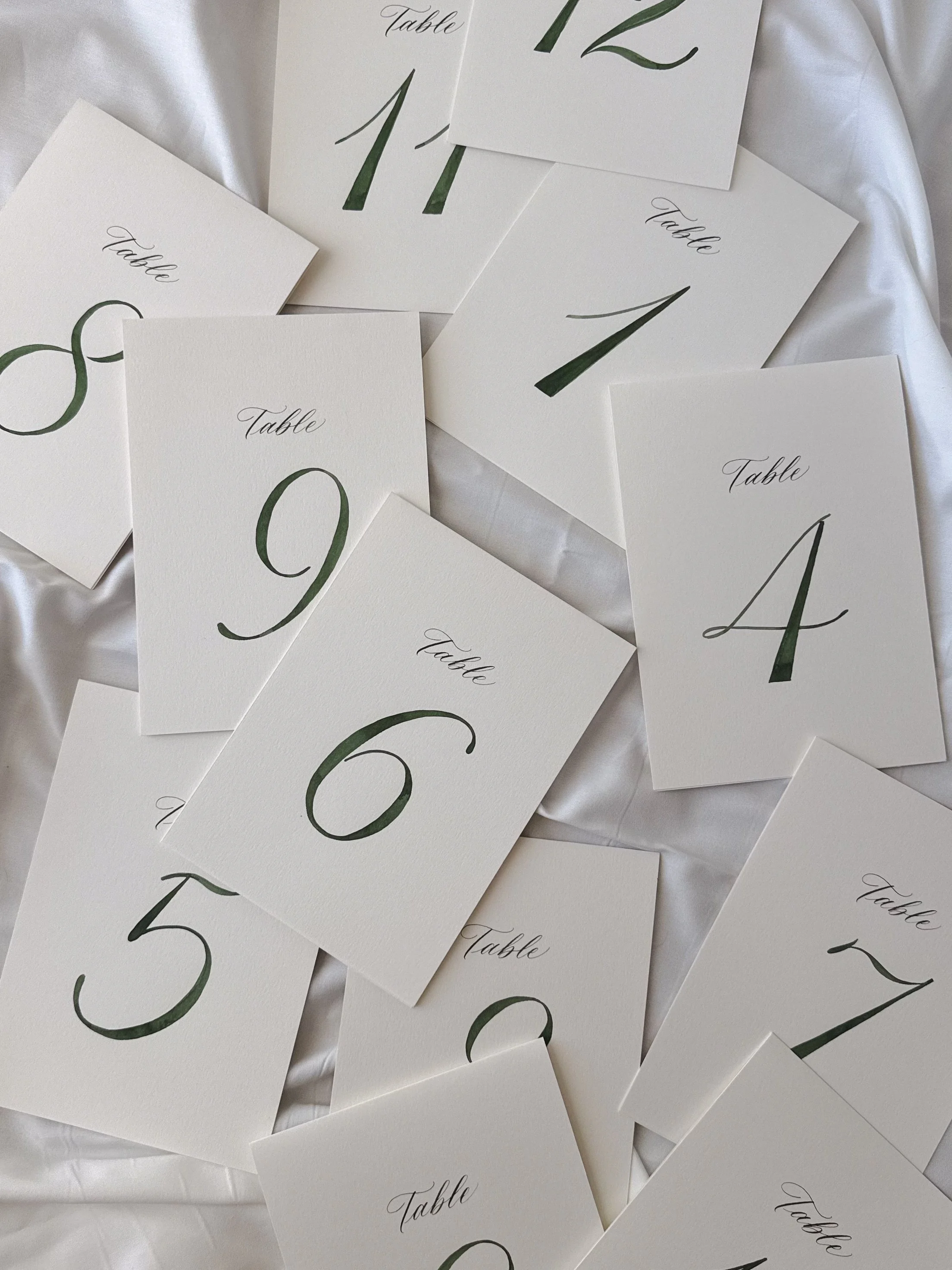 Multiple white table cards with green handwritten numbers and the word 'Table' in cursive, scattered on a white fabric surface.