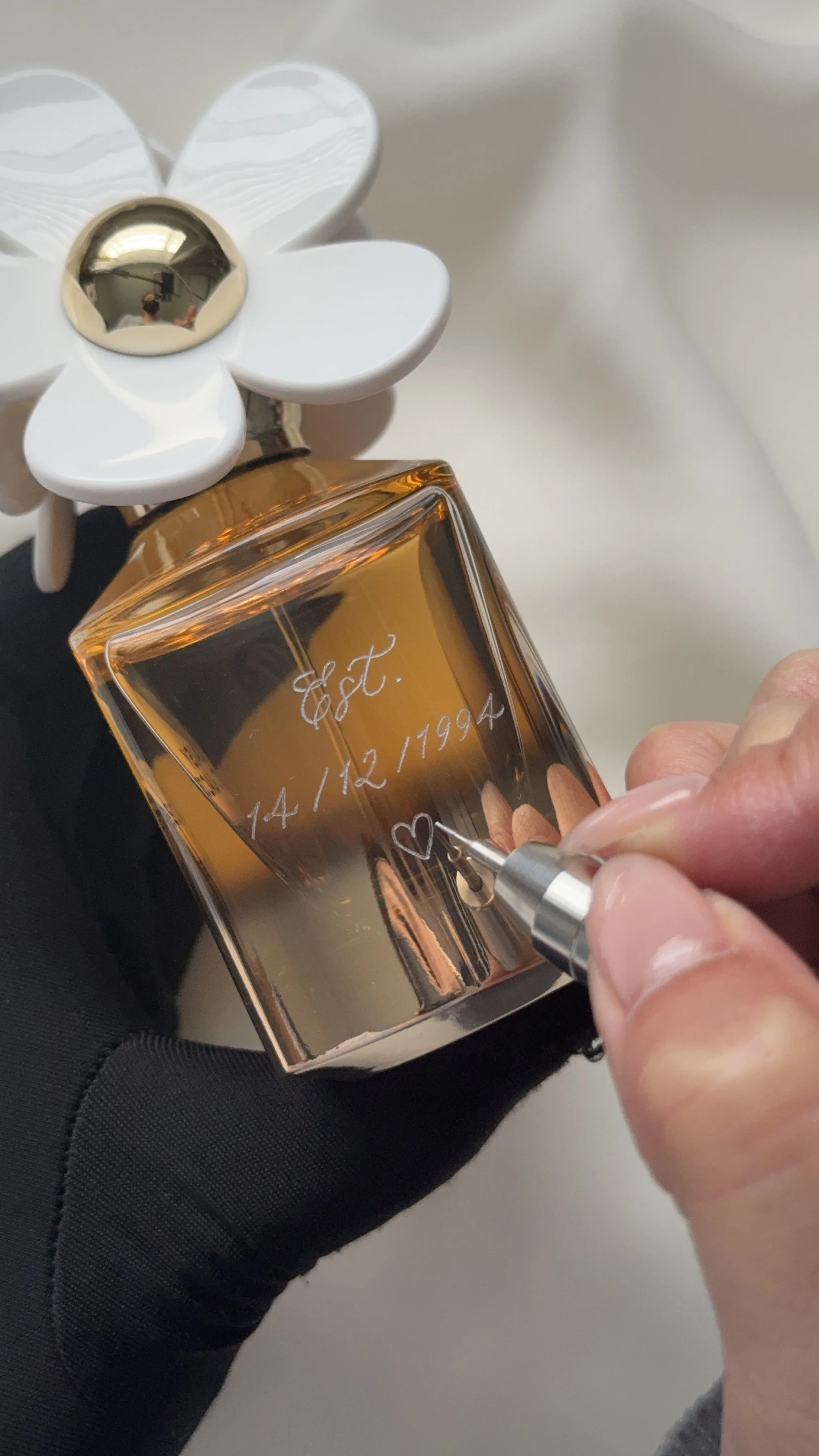 A hand holding a pen is engraving a heart and a date, '14/12/1994,' on a gold-colored perfume bottle. A white flower-shaped decoration with a gold center is attached to the top of the bottle. The person is wearing a black glove.