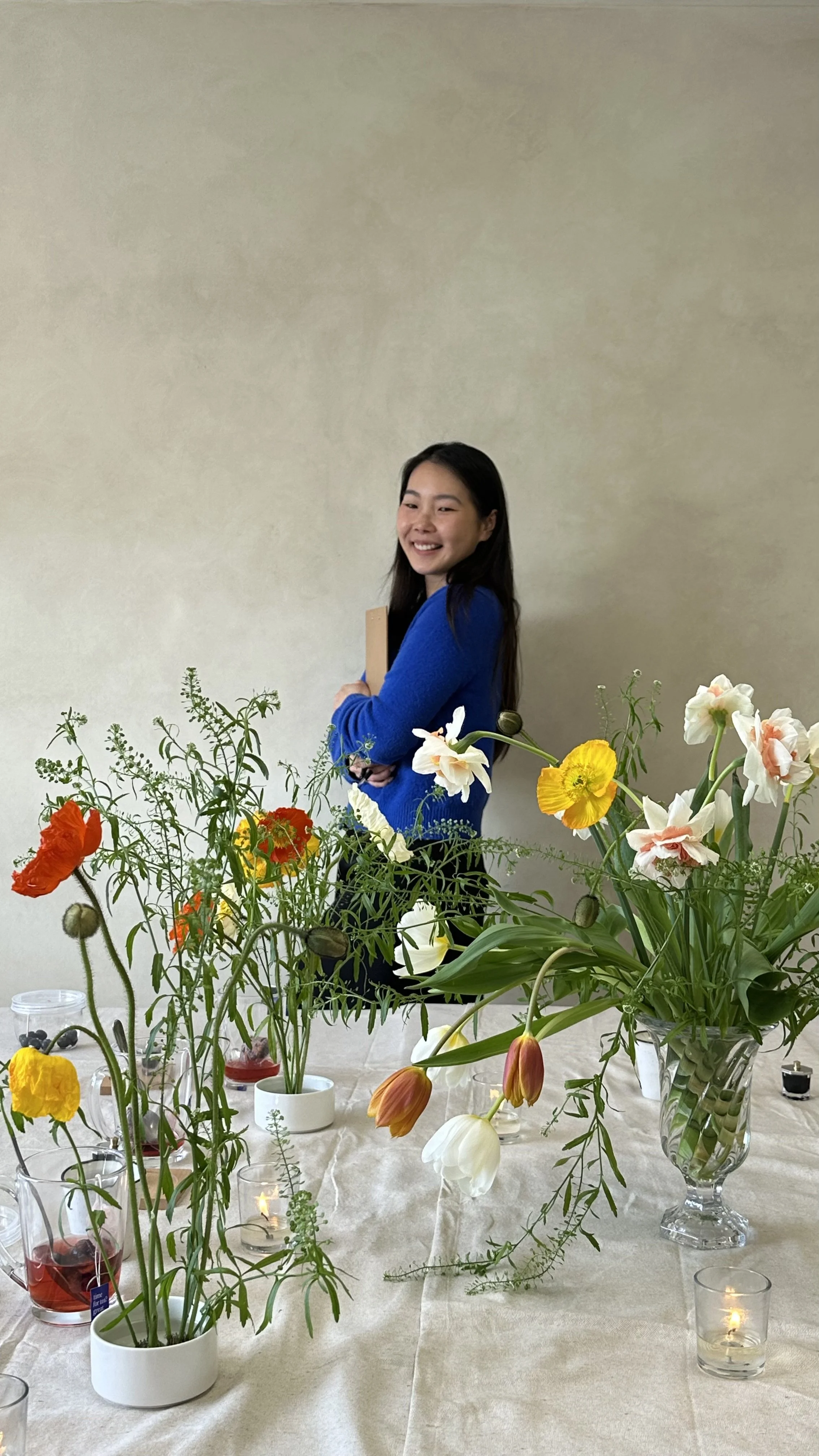 A young woman with long dark hair smiling and standing behind a table with an assortment of colorful flowers in various vases and small candles.