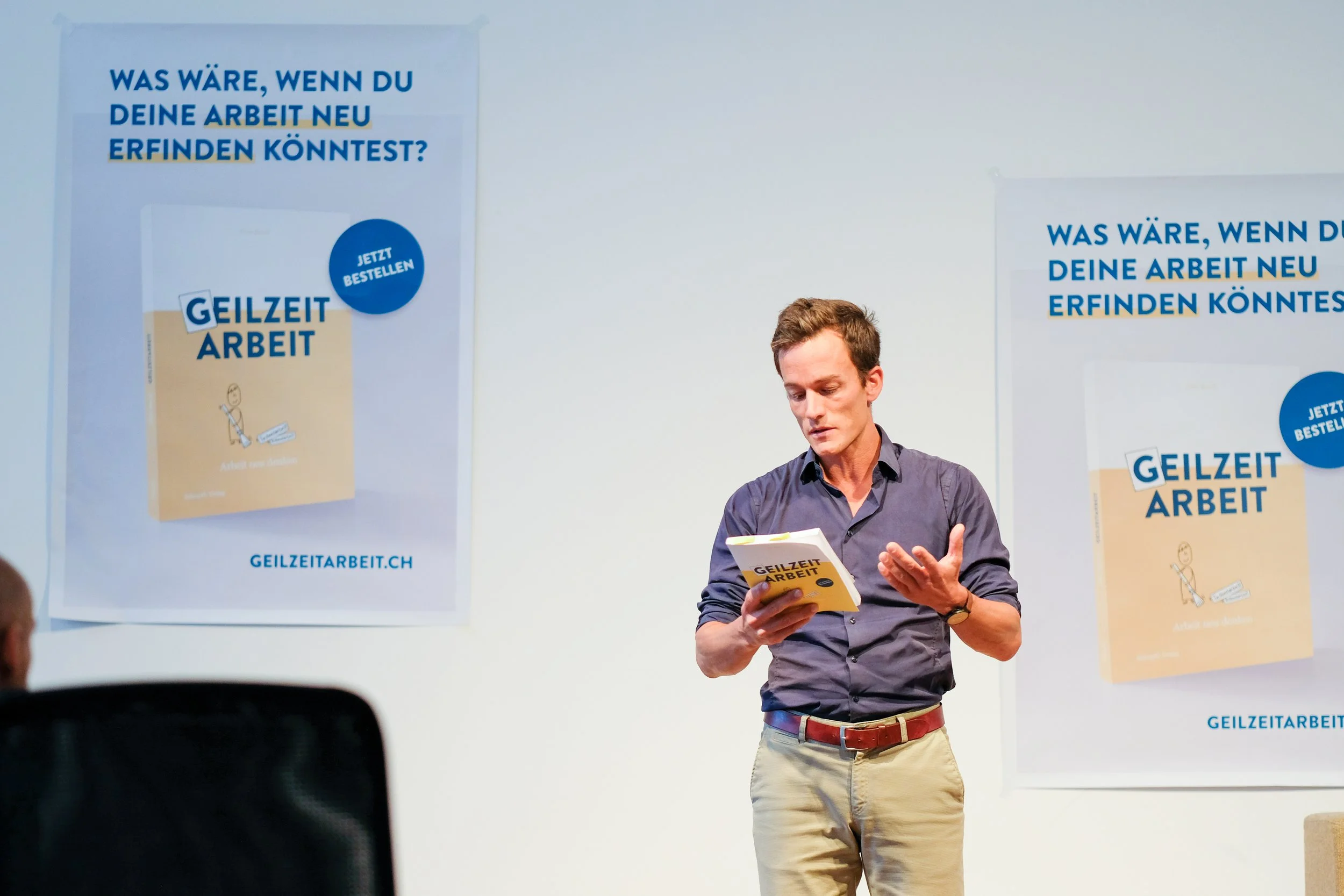 Ein Mann liest ein Buch mit dem Titel "Geilzeit Arbeit" bei einer Präsentation, im Hintergrund Plakate mit dem gleichen Buchtitel und einem Slogan in deutscher Sprache.
