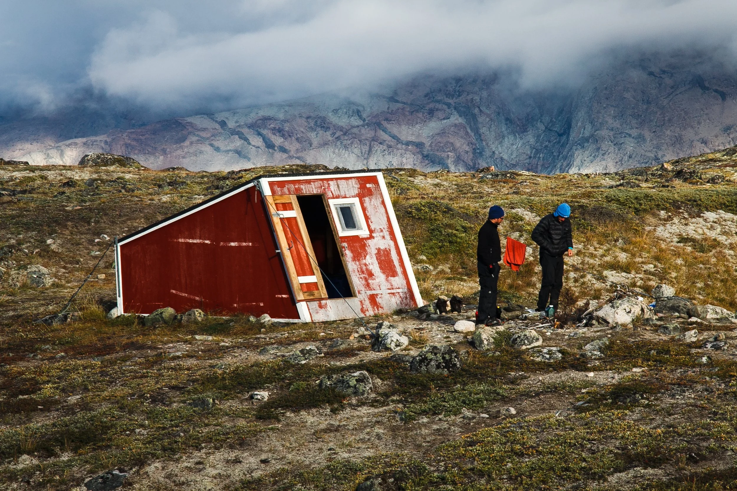 Zwei Menschen stehen vor einem umgestürtzen roten Hüttendach in einer kargen, felsigen Landschaft mit Bergen im Hintergrund und dunklen Wolken am Himmel.