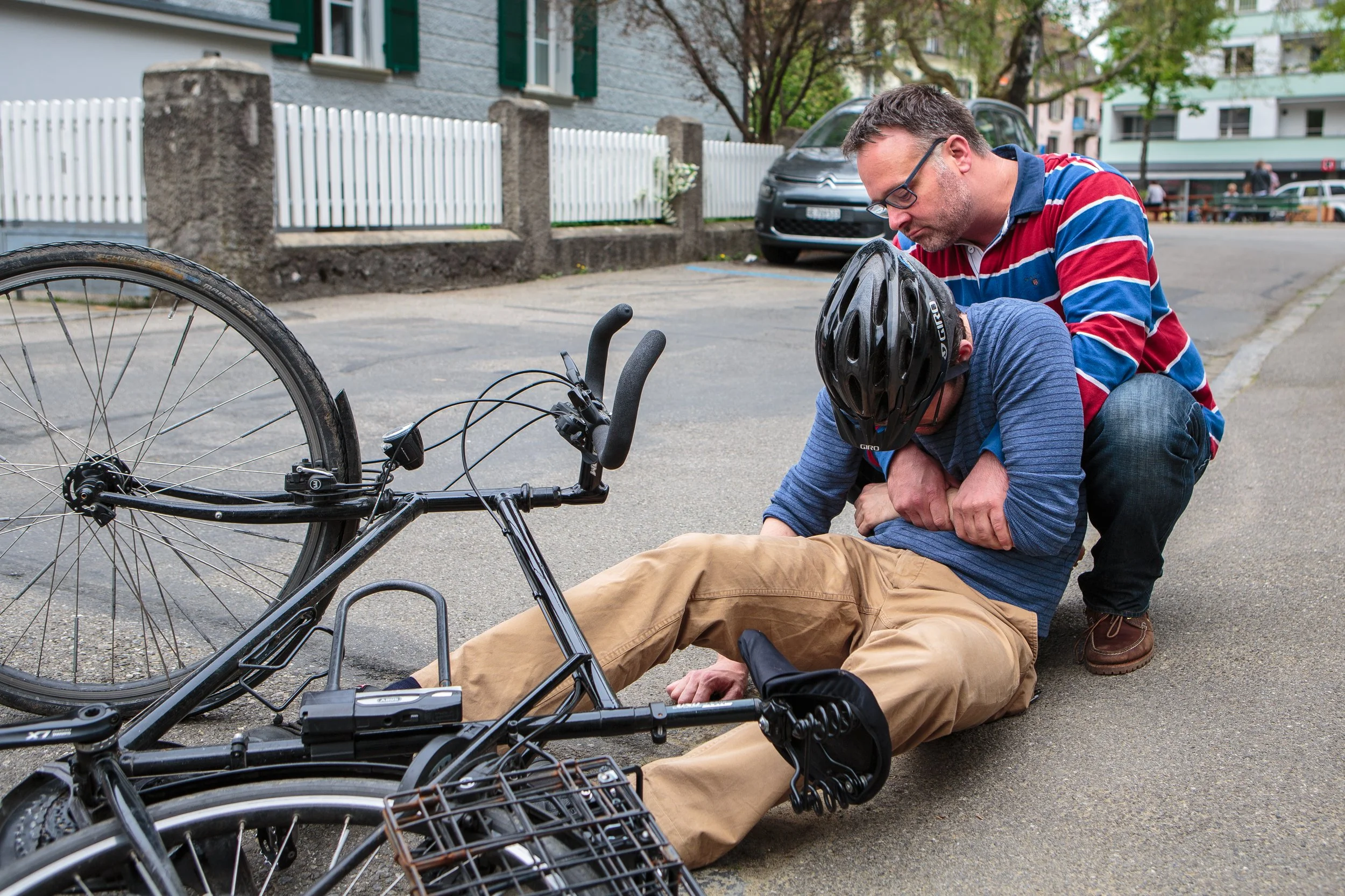 Ein Mann liegt verletzt auf der Straße nach einem Fahrradunfall, während ein weiterer Mann ihm hilft, ihn zu stabilisieren.