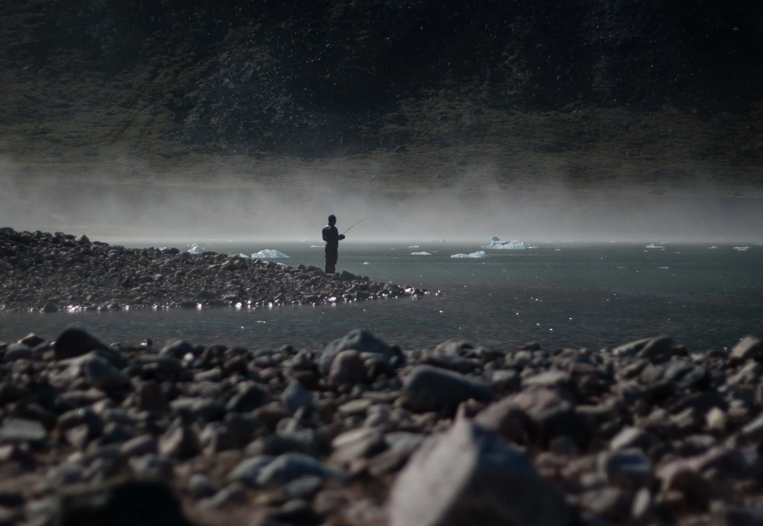 Ein einsamer Angler steht an einem gletscherbedeckten Fluss in einer kalten, bergigen Landschaft, umgeben von Steinen und Eisbergen.