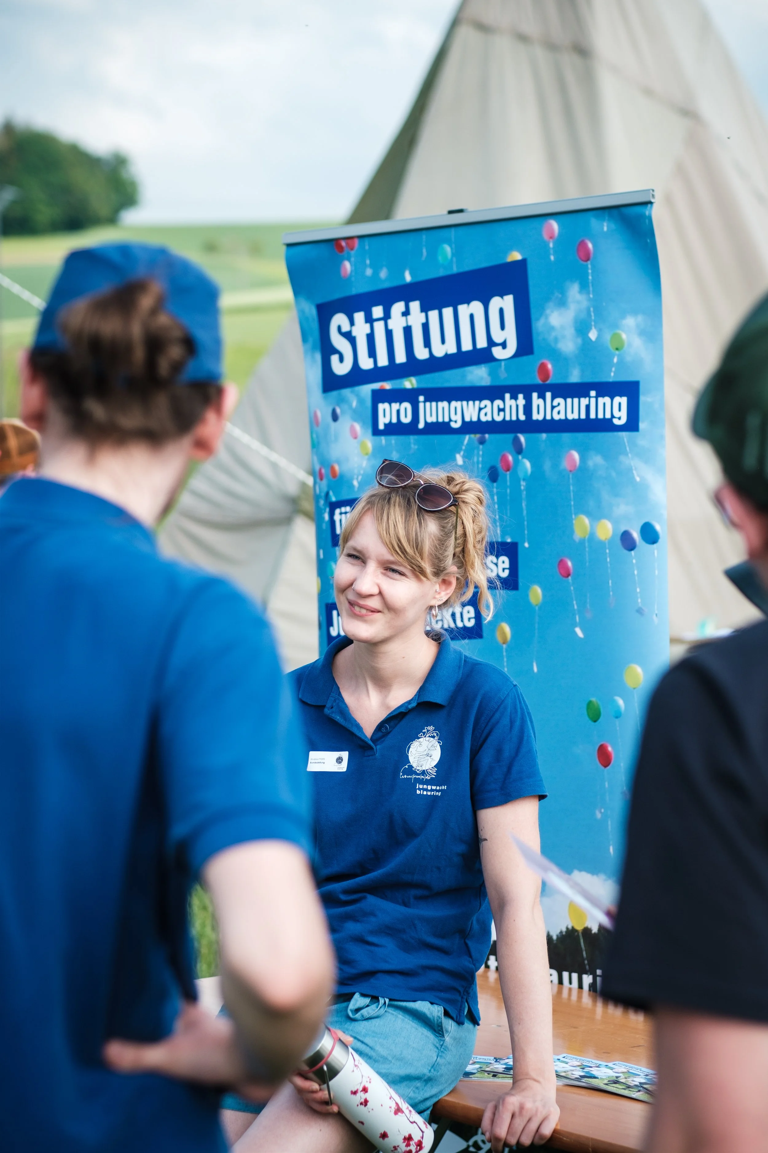 Eine junge Frau spricht mit zwei Männern an einem Stand bei einem Outdoor-Event, im Hintergrund ein Banner mit der Aufschrift 'Stiftung pro Jungwacht blauring' und bunten Ballons, auf einer Wiese mit Zelten im Freien.