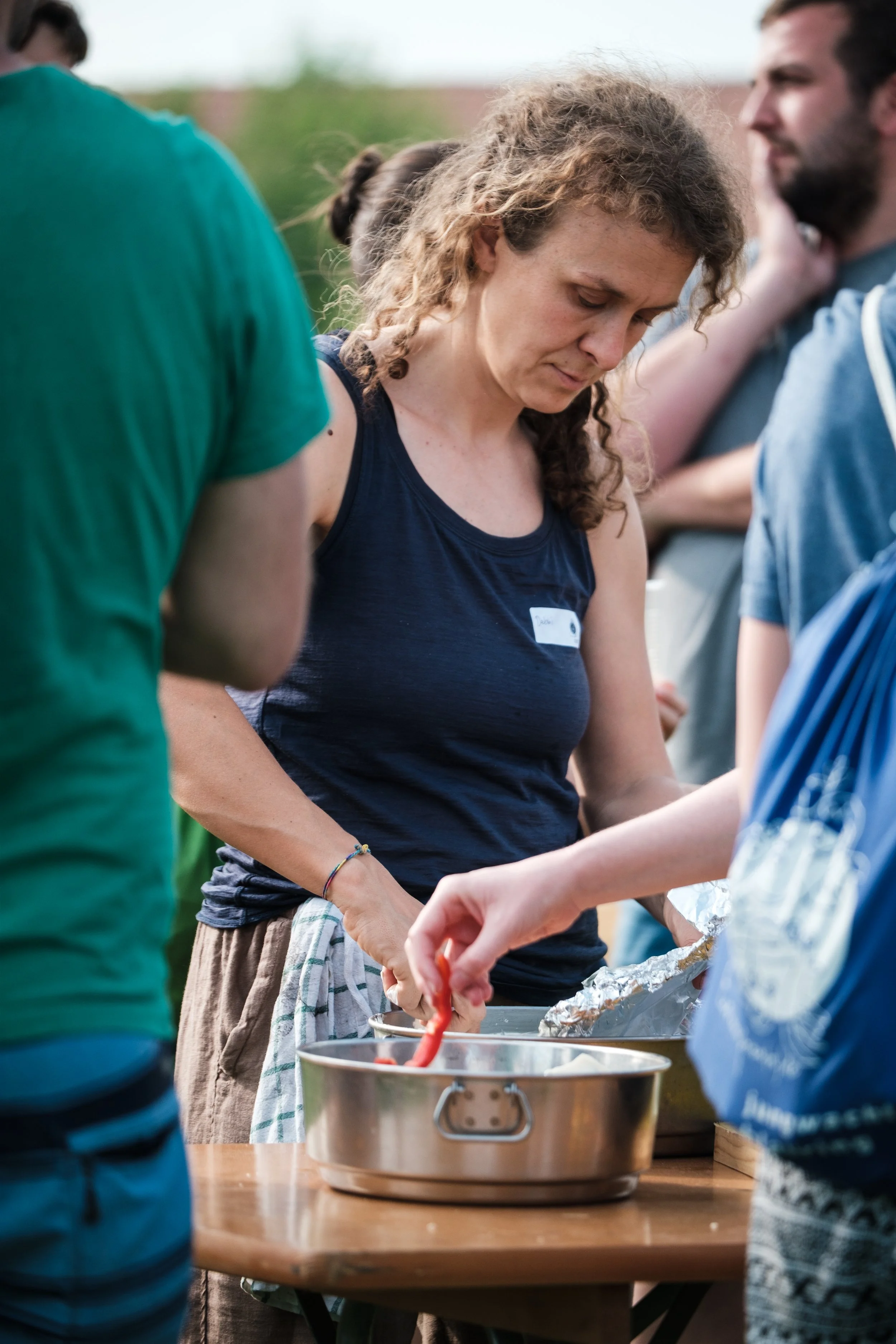 Menschen bei einem Outdoor-Grillabend bereiten Essen zu, Woman in blauer top, umgeben von anderen Personen, mit Grillzubehör auf einem Holztisch.