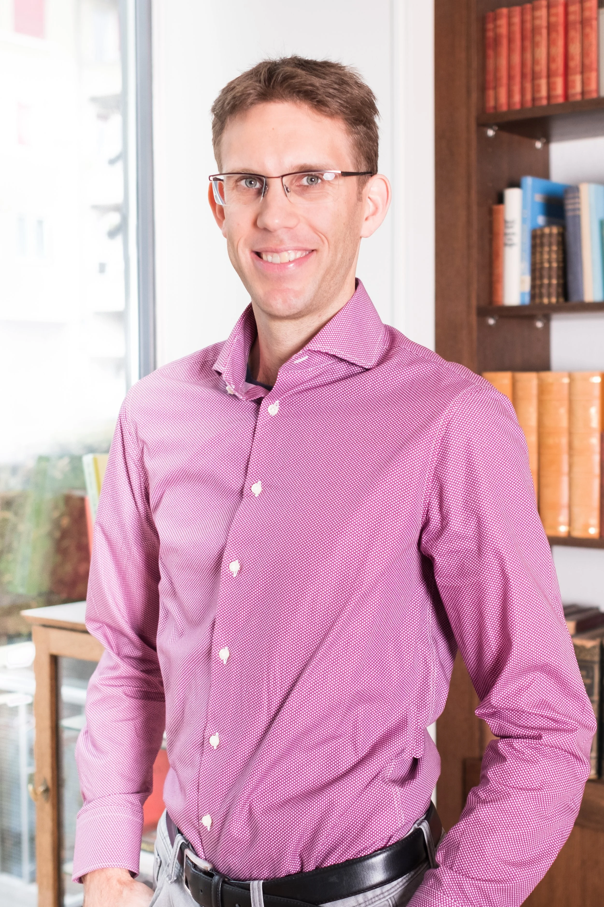 Ein Mann mit Brille, im Büro, trägt ein pinkfarbenes Hemd und lächelt.