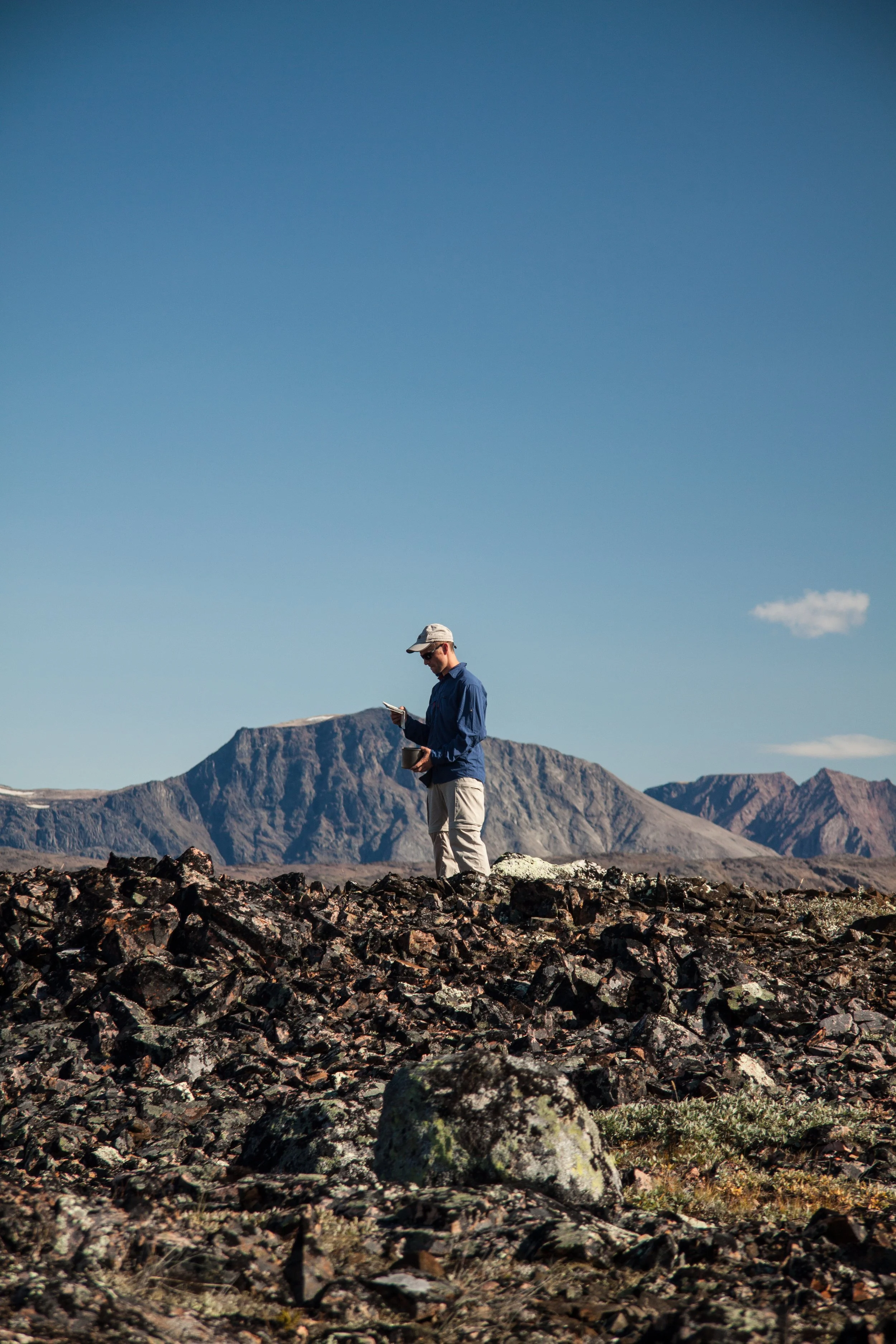 Ein Mann steht in einer felsigen, wüstenähnlichen Landschaft mit Bergen im Hintergrund und liest in einem Buch.