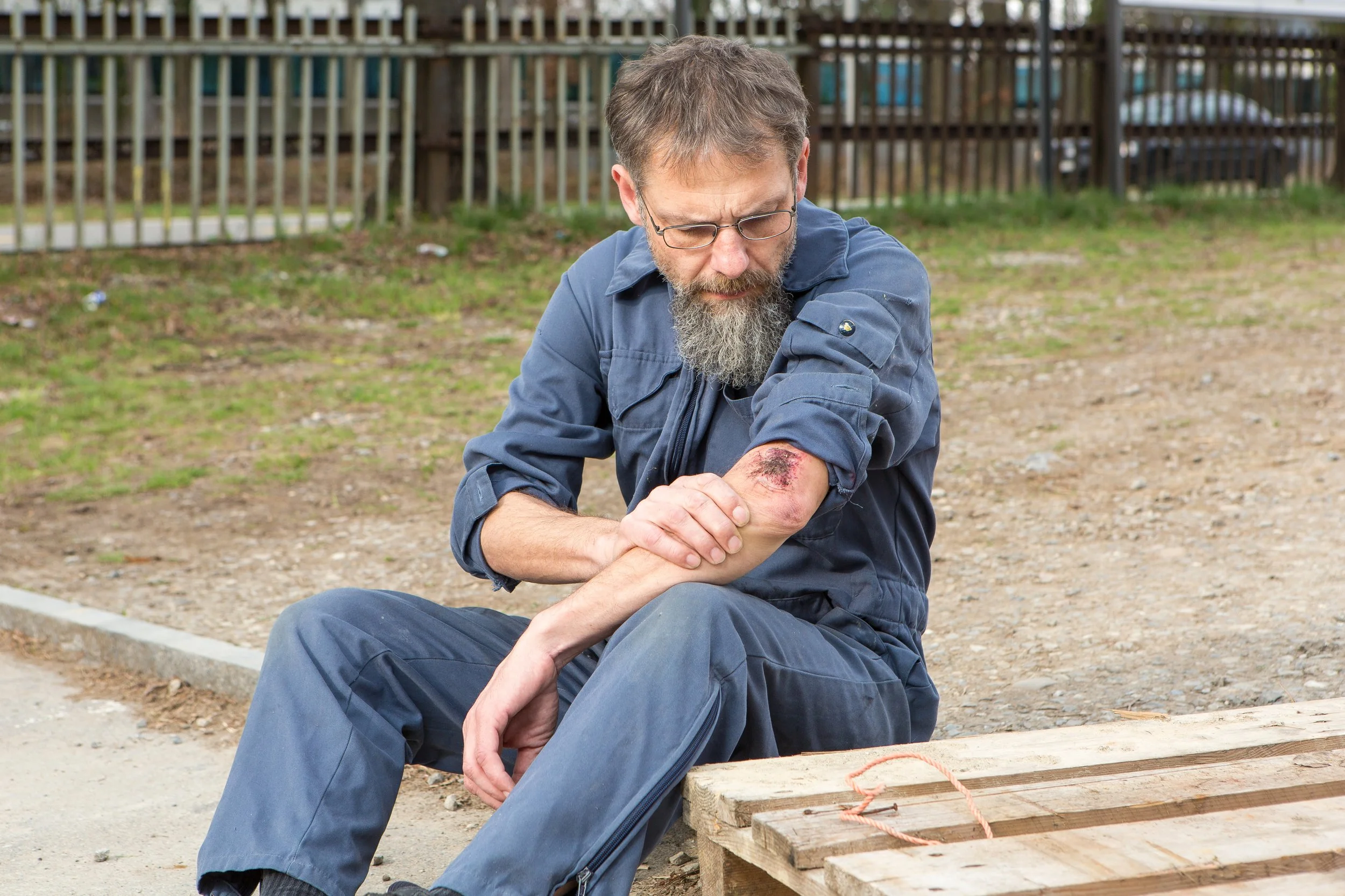 Ein Mann mit Brille und Bart sitzt auf dem Boden im Freien und betrachtet eine Verletzung an seinem Arm, die blutig und geschwollen ist.