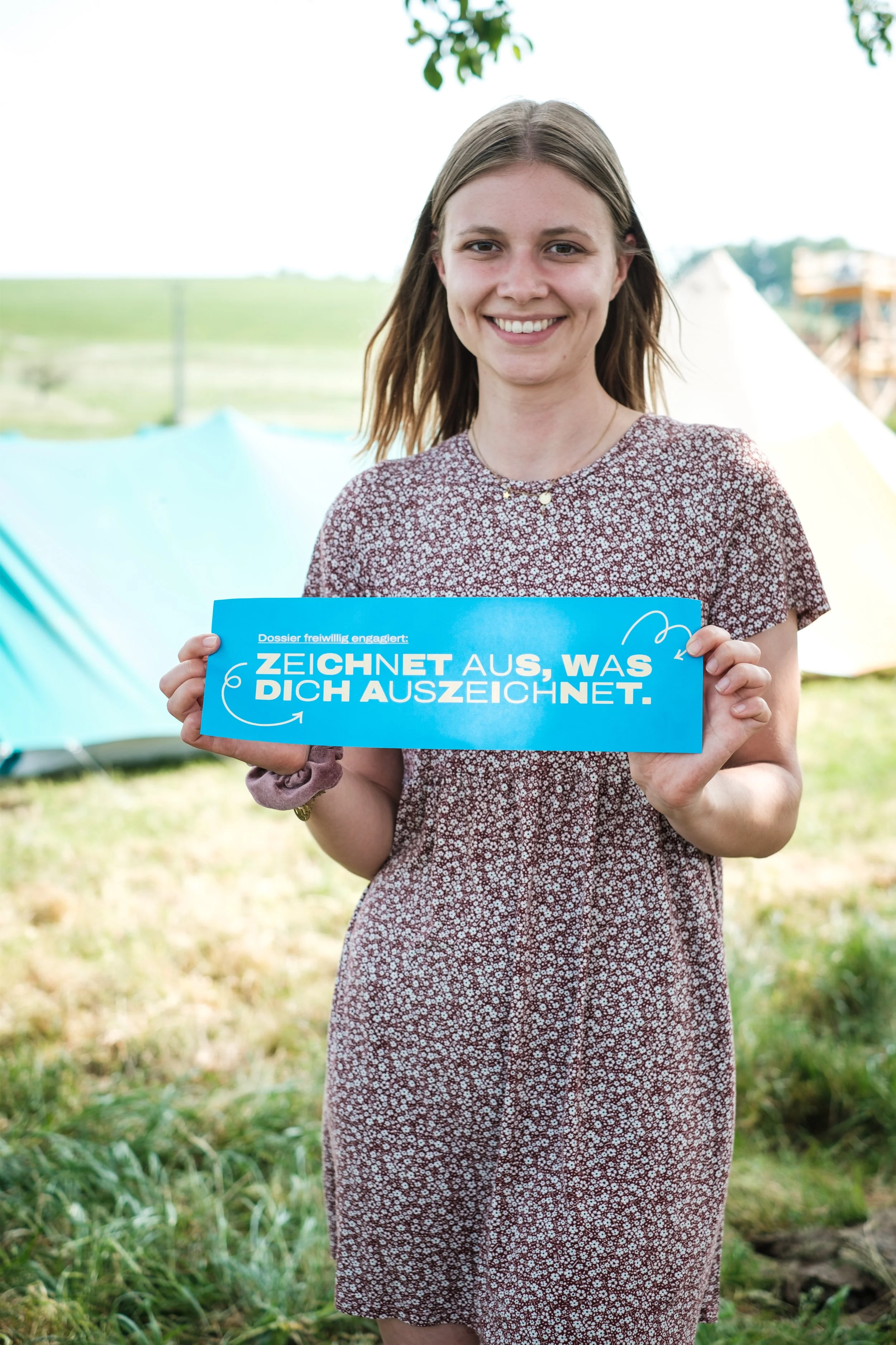 Junge Frau im Freien hält ein blaues Schild mit der Aufschrift 'Zeichnet aus, was dich auszeichnet'.
