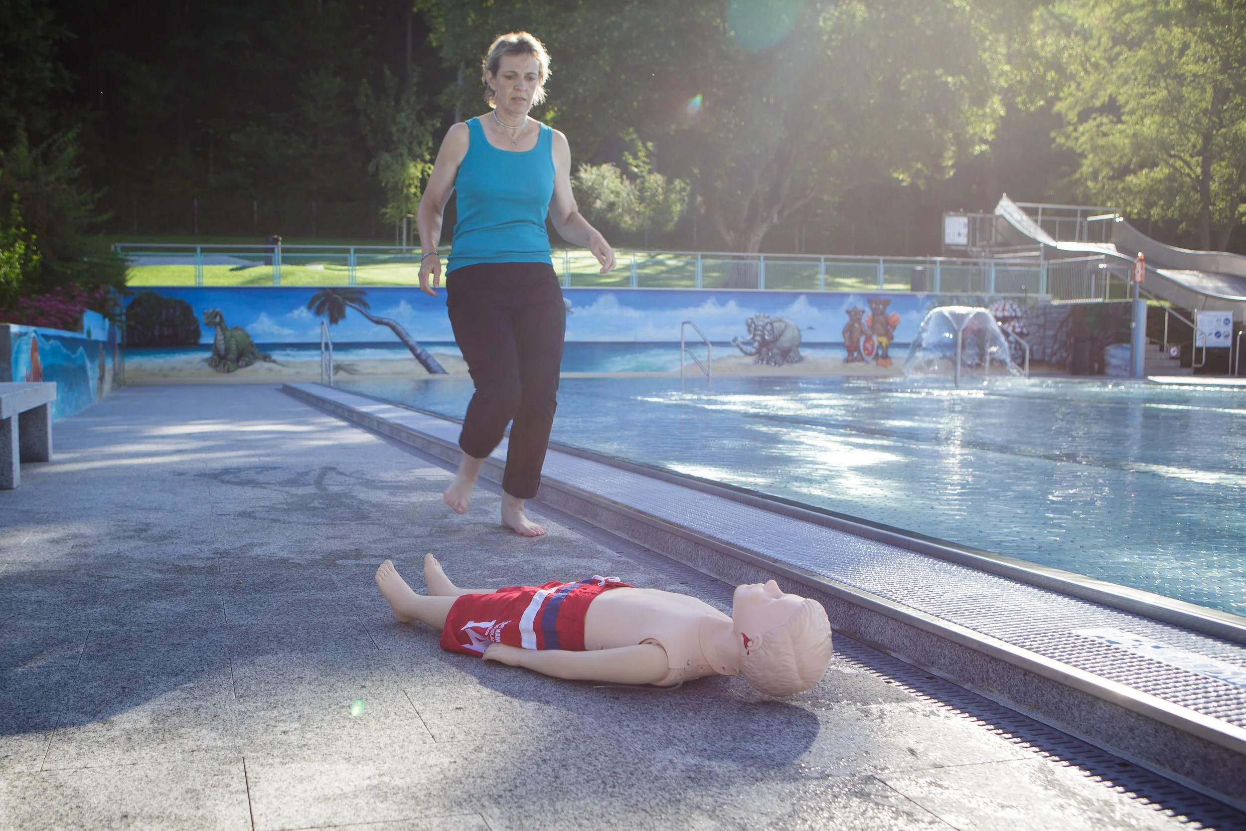 Eine ältere Frau steht neben einem auf dem Boden liegenden dummy und übt Erste Hilfe an einem städtischen Swimmingpool.