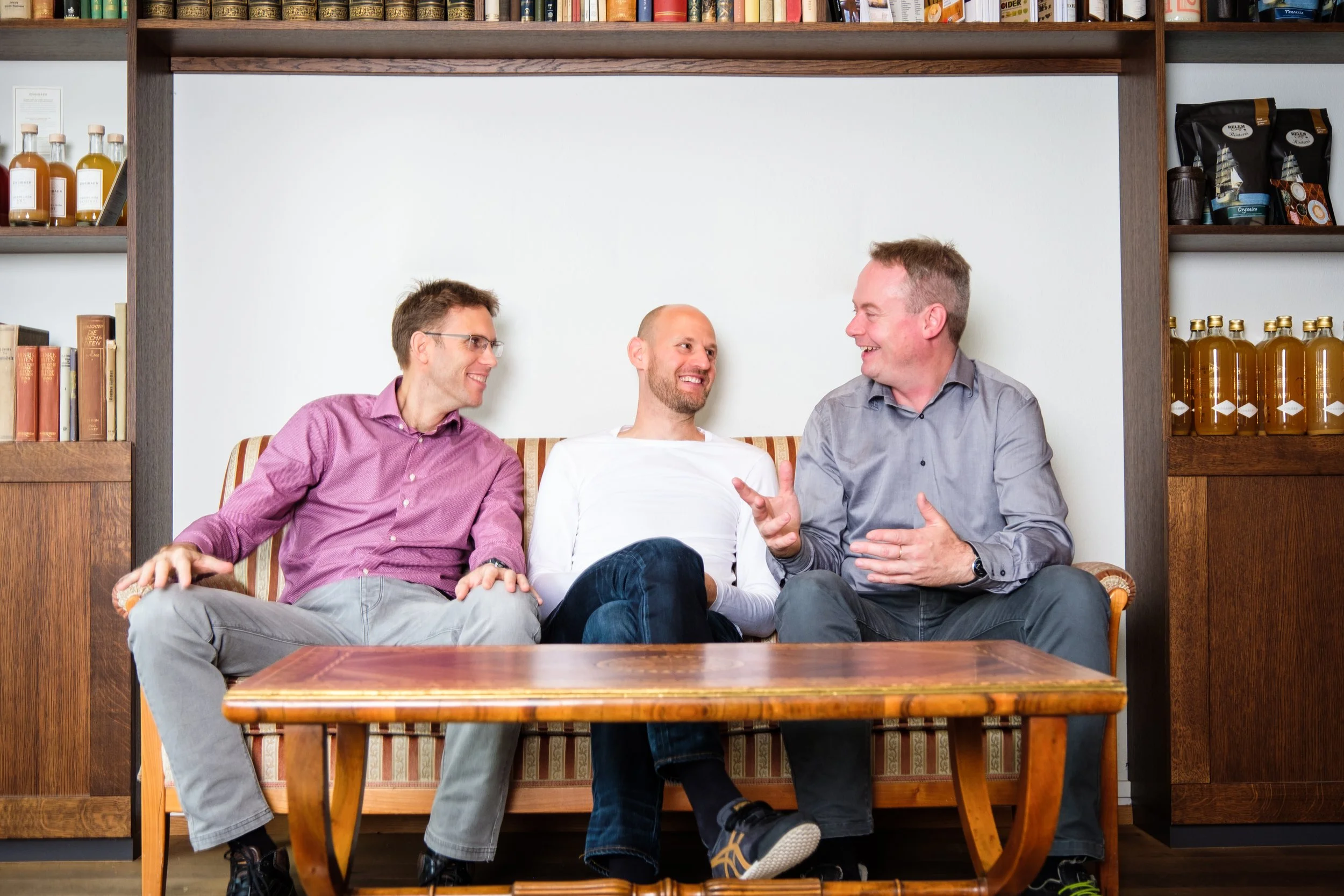 Drei Männer sitzen auf einem Sofa und unterhalten sich lachend in einem Wohnzimmer mit Büchern und Flaschen im Hintergrund.