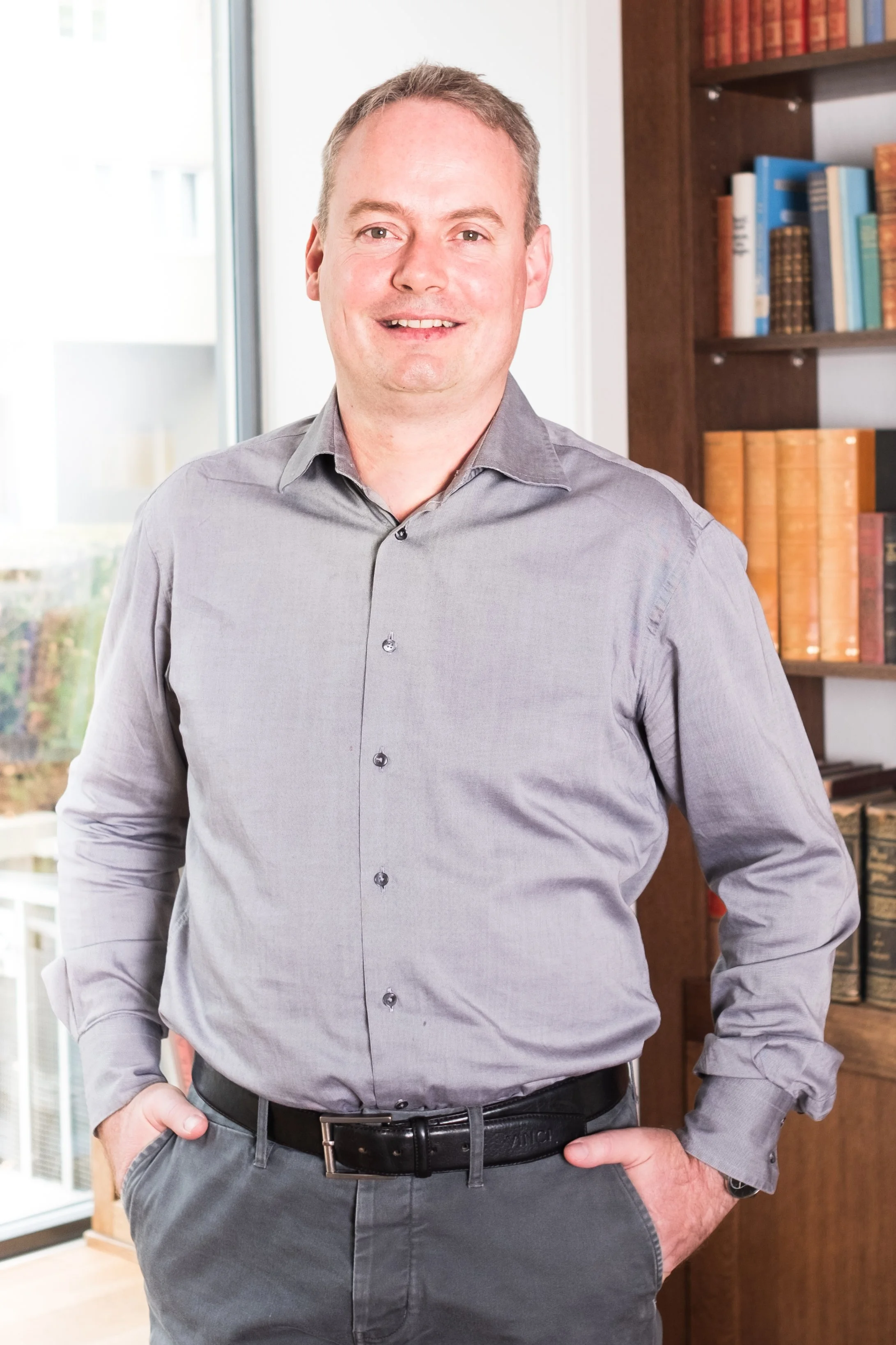 Ein Mann in einem grauen Hemd steht in einem Raum mit Bücherregalen im Hintergrund.