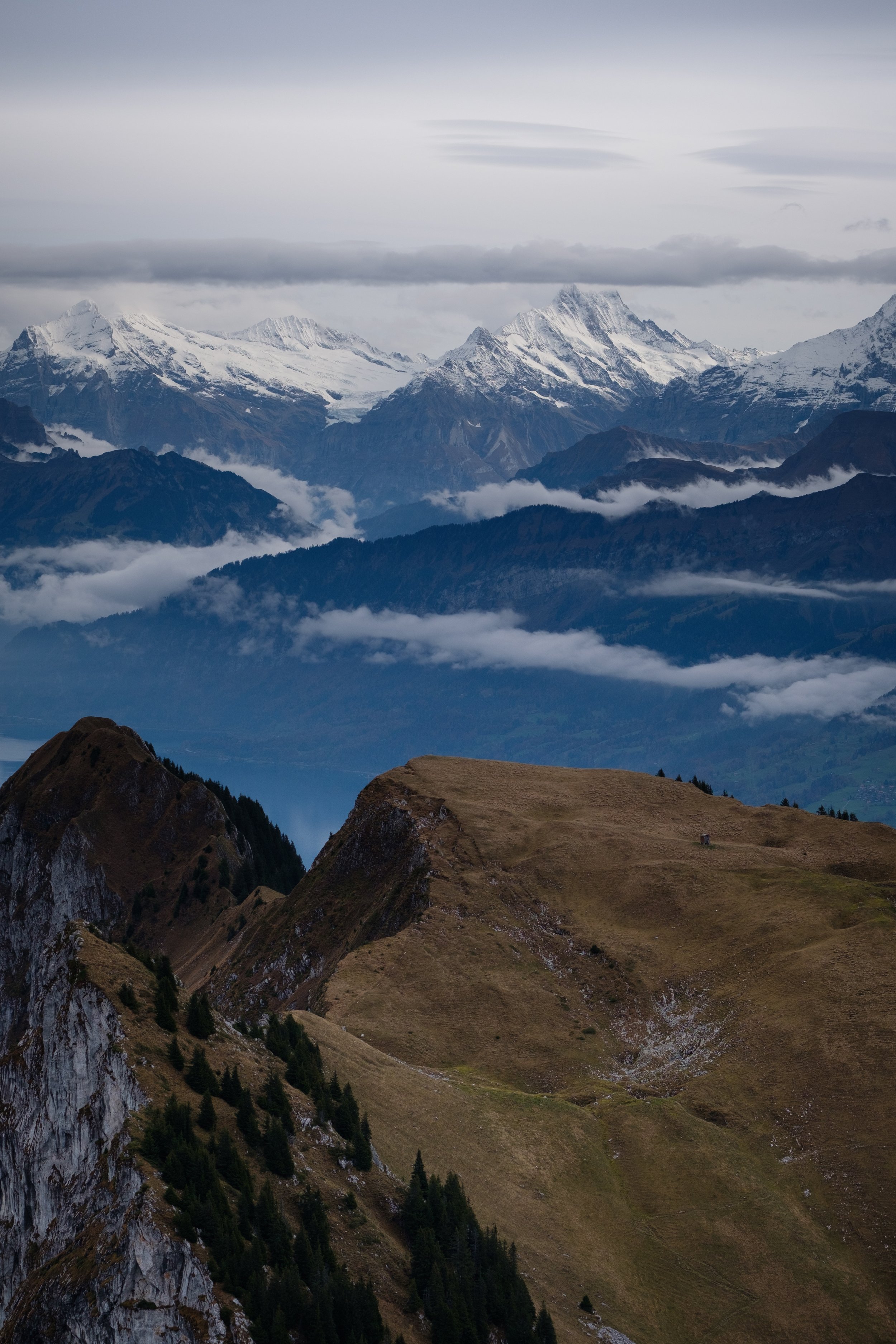 Berglandschaft mit schneebedeckten Gipfeln im Hintergrund, Wolken, Täler und grasbewachsene Hügel im Vordergrund.