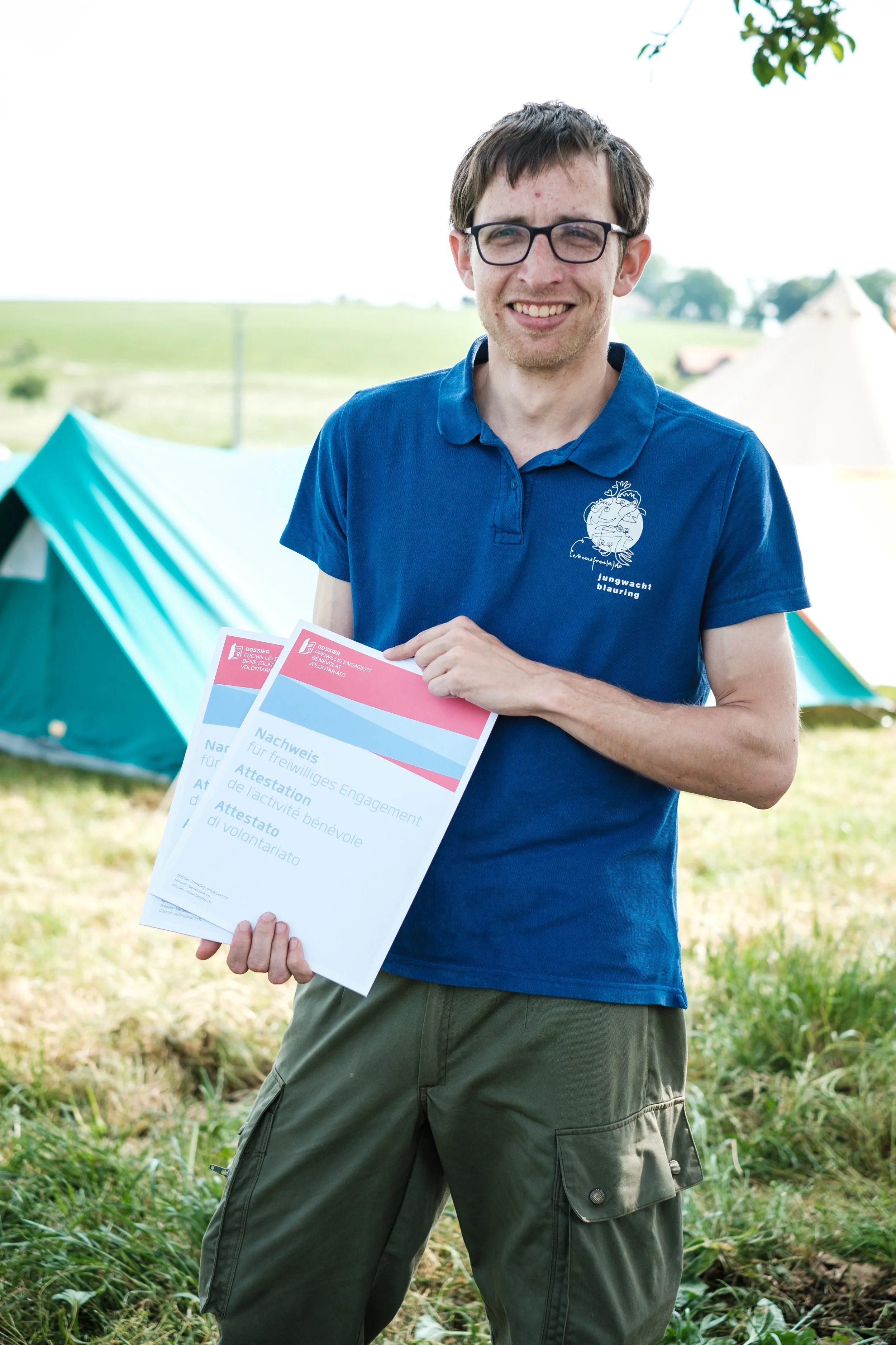 Junger Mann mit Brille hält Zertifikate für Freiwilliges Engagement in die Kamera, im Freien mit Zelten im Hintergrund.