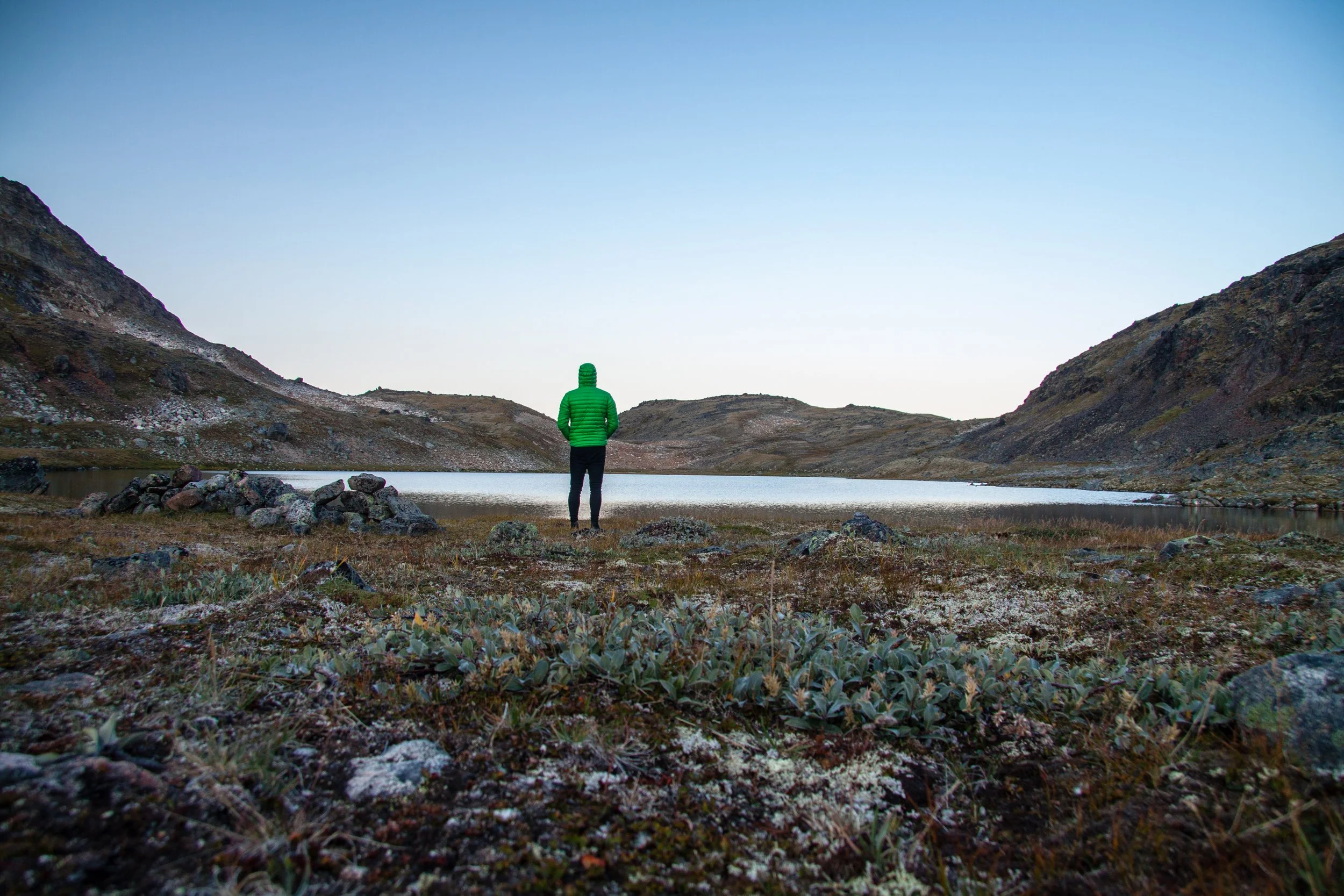 Eine einsame Person in grüner Jacke steht vor einem See in einer bergigen, kargen Landschaft bei Sonnenuntergang.