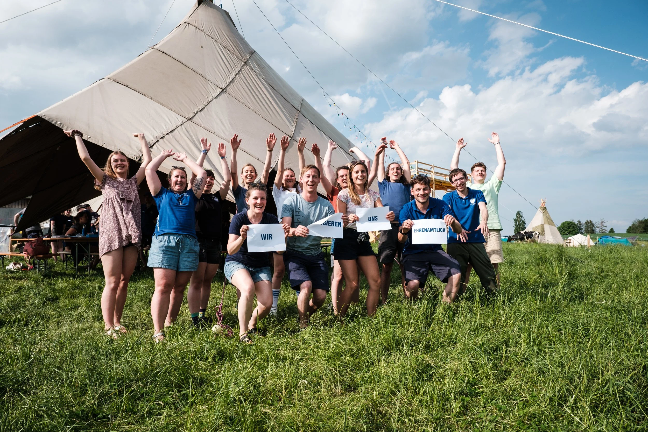 Gruppe von jungen Menschen, die auf einem Feld mit Zelten im Hintergrund stehen und schwarz-weiße Schilder mit deutschen Worten halten, die zusammen den Satz bilden: "Wir engagieren uns ehrenamtlich". Sie lachen und haben die Arme in die Luft geworfe