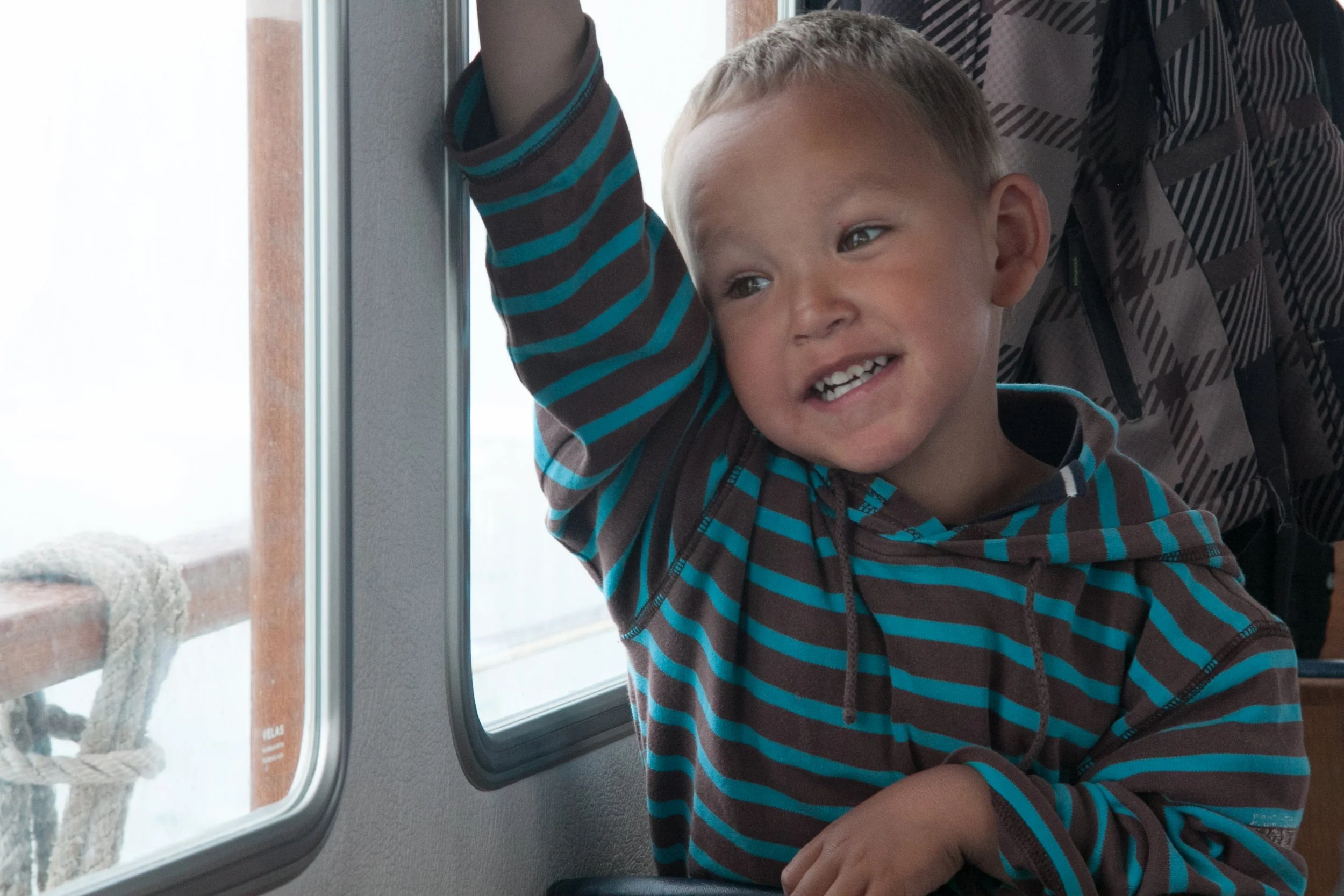 Ein lachender Junge mit kurzen blondem Haar, der in einem Bus oder Zug an einem Fenster sitzt, trägt ein gestreiftes Hoodie in Blau, Grau und Schwarz, und hält die Hand an die Scheibe.