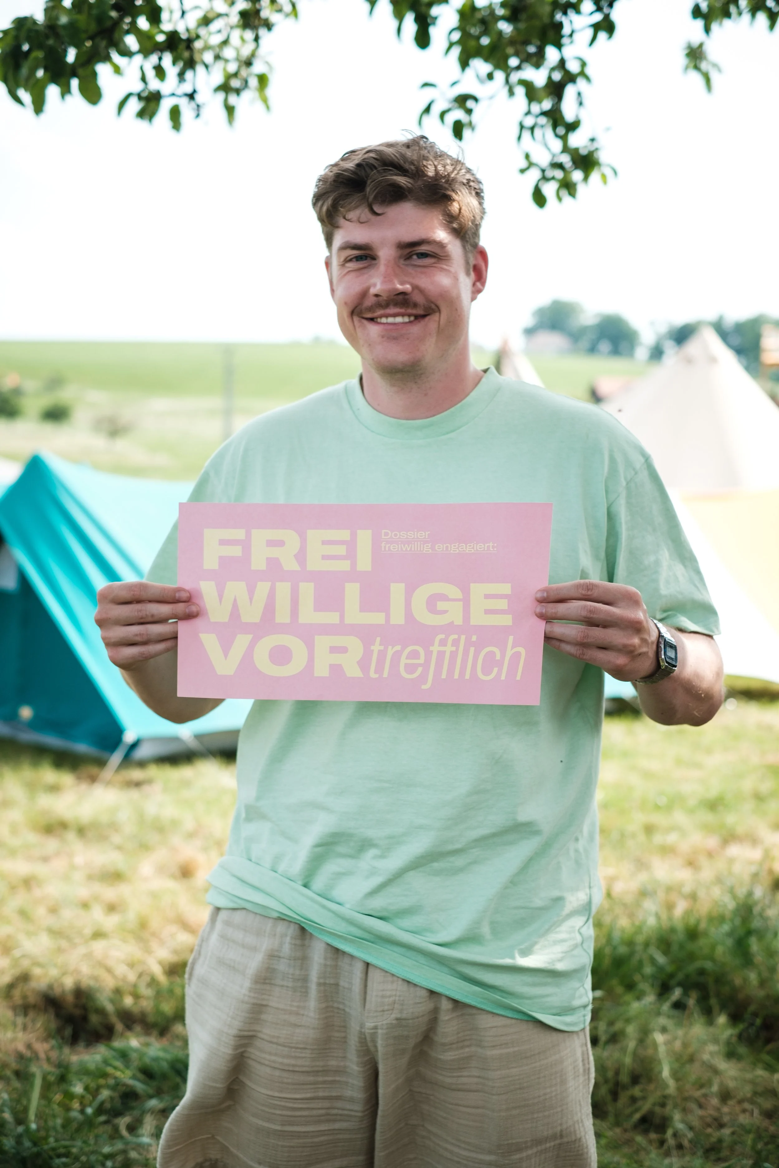 Ein lachender Mann hält ein pinkfarbenes Schild mit dem deutschen Text „FREI WILLIGE VOR trefflich“ bei einem Campingplatz im Freien, im Hintergrund sind grüne Wiesen, Bäume und Zelte.