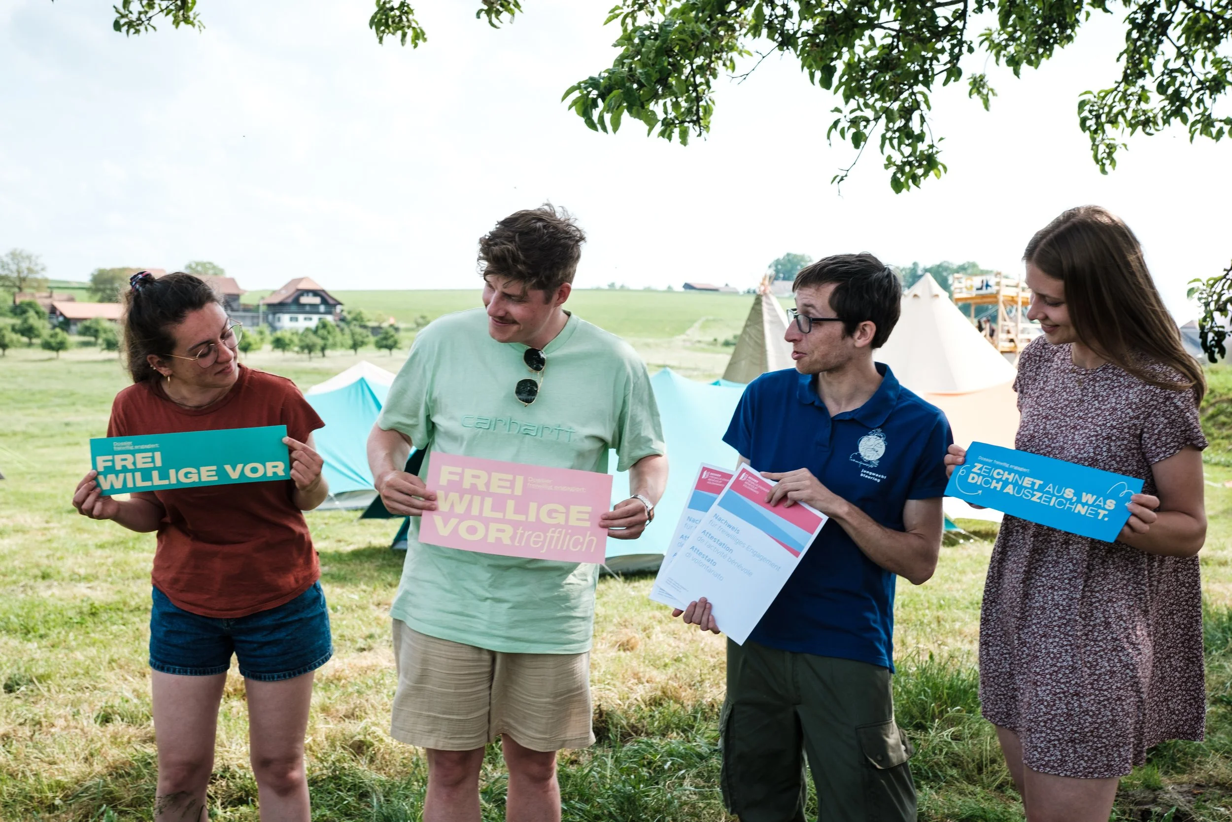 Vier Menschen stehen im Freien unter einem Baum und halten Plakate mit Texten. Sie scheinen in einer entspannten, freundlichen Diskussion zu sein, mit Zelten im Hintergrund und offenen Feldern bei schönem Wetter.