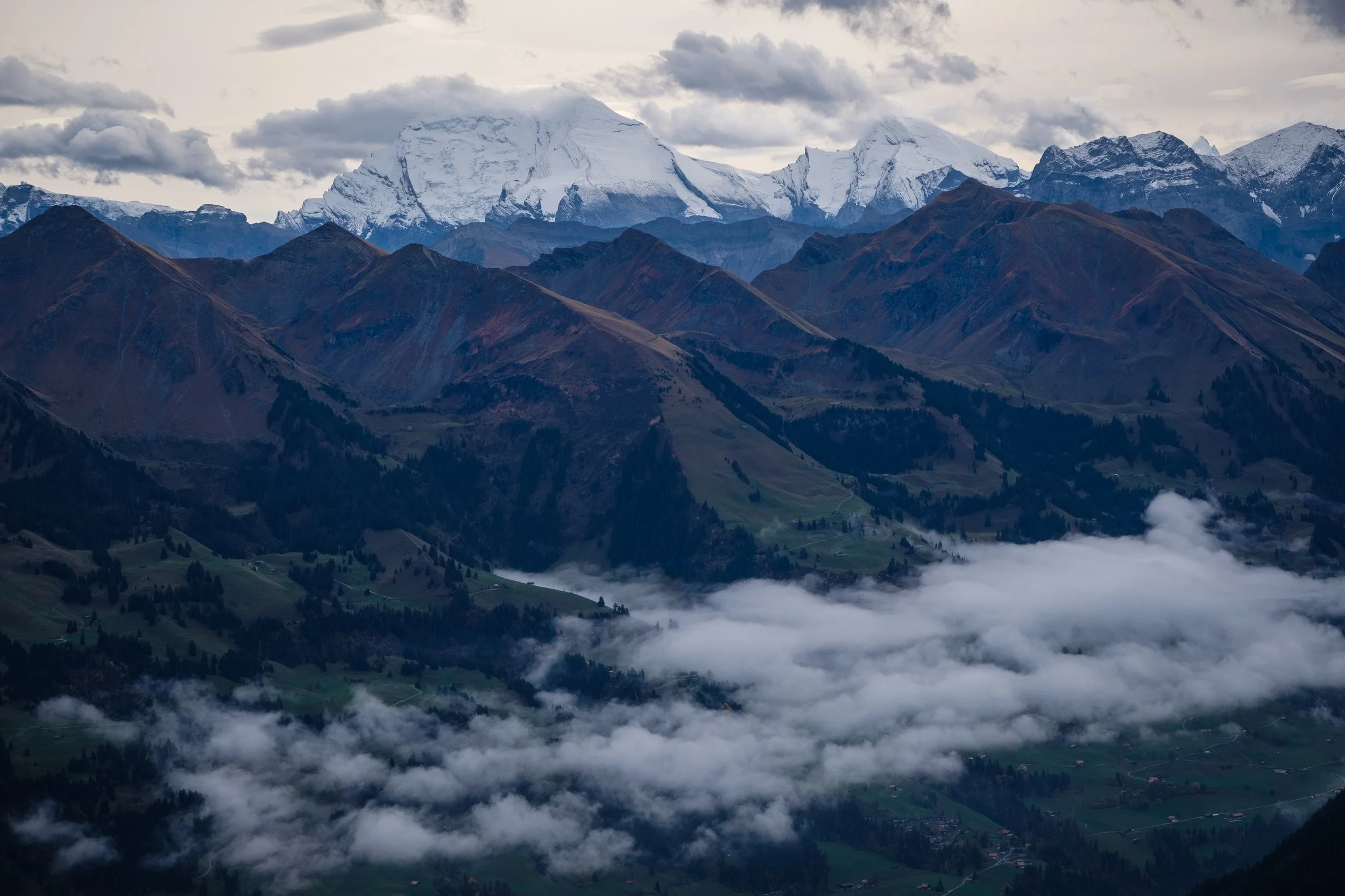 Berglandschaft mit schneebedecktem Gipfel im Hintergrund, bewaldeten Hügeln und Wolken im Tal.