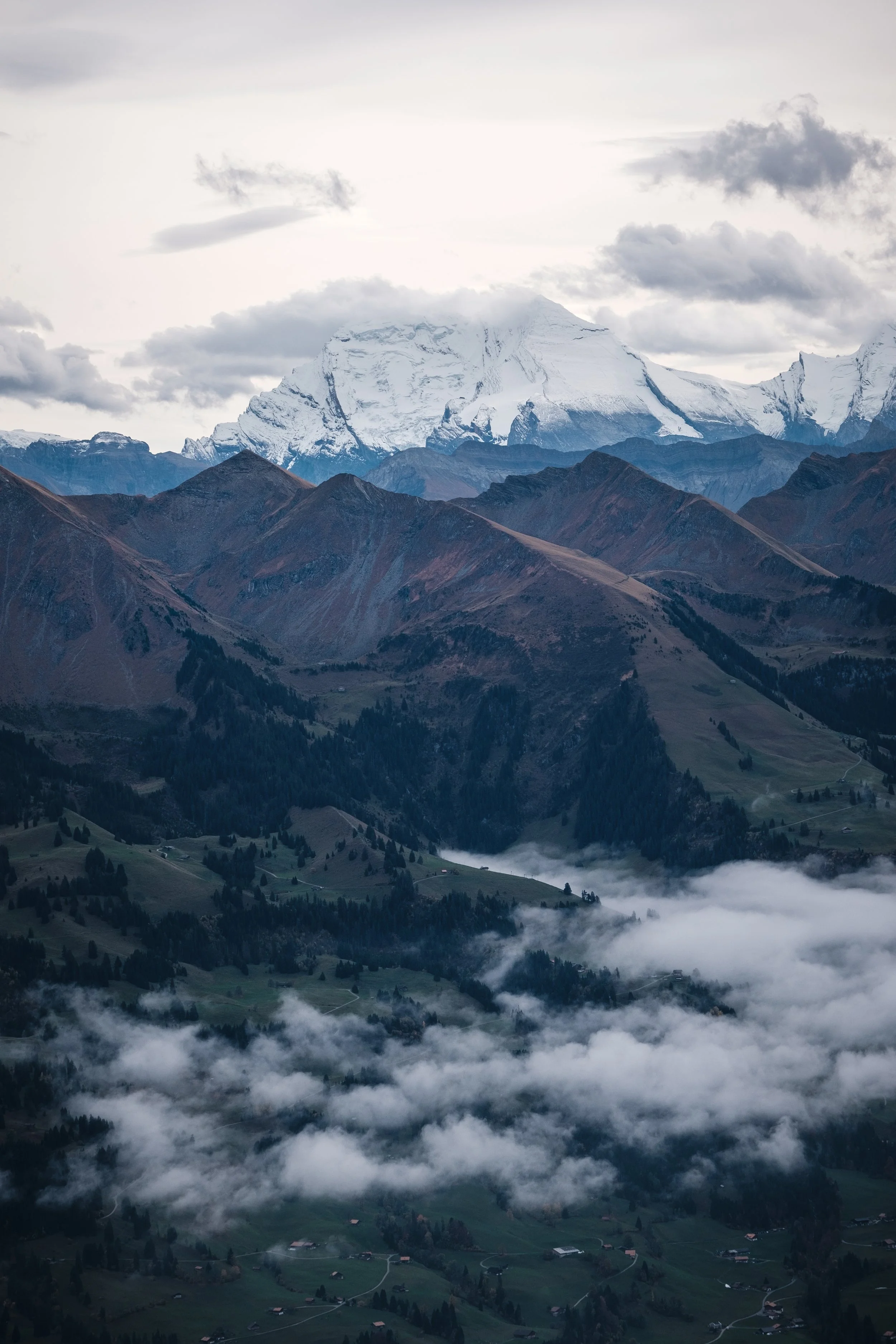 Berglandschaft mit schneebedecktem Gipfel im Hintergrund, grüne Hügel und windige Wolken im Vordergrund.