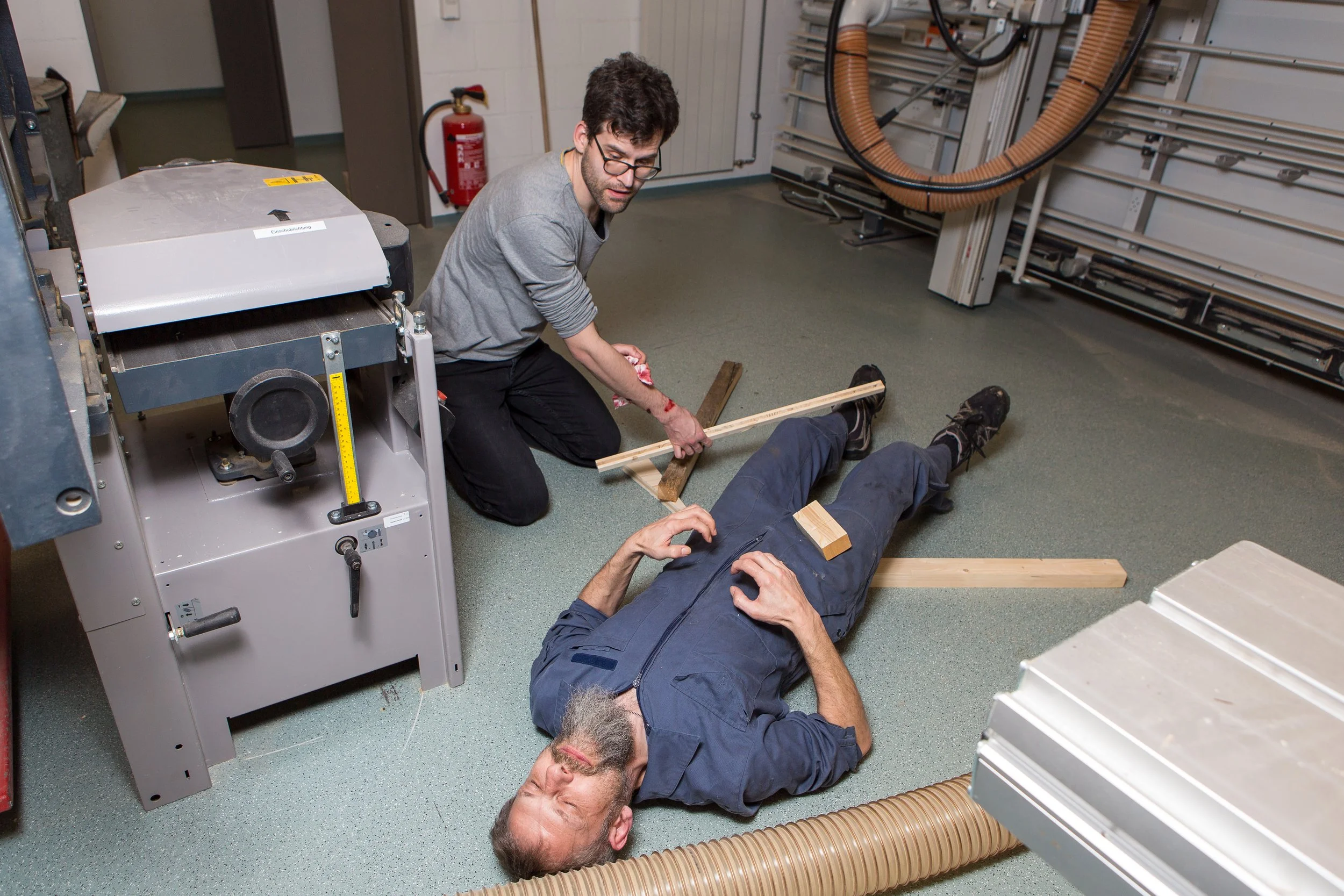 Zwei Männer in einer Werkstatt, einer liegt auf dem Boden mit einem Holzstück auf dem Bauch, der andere sitzt daneben, mit einem Bluterguss am Arm, und hält ein langes Holzstück. Es sieht aus wie eine Notfallsituation oder Unfall.