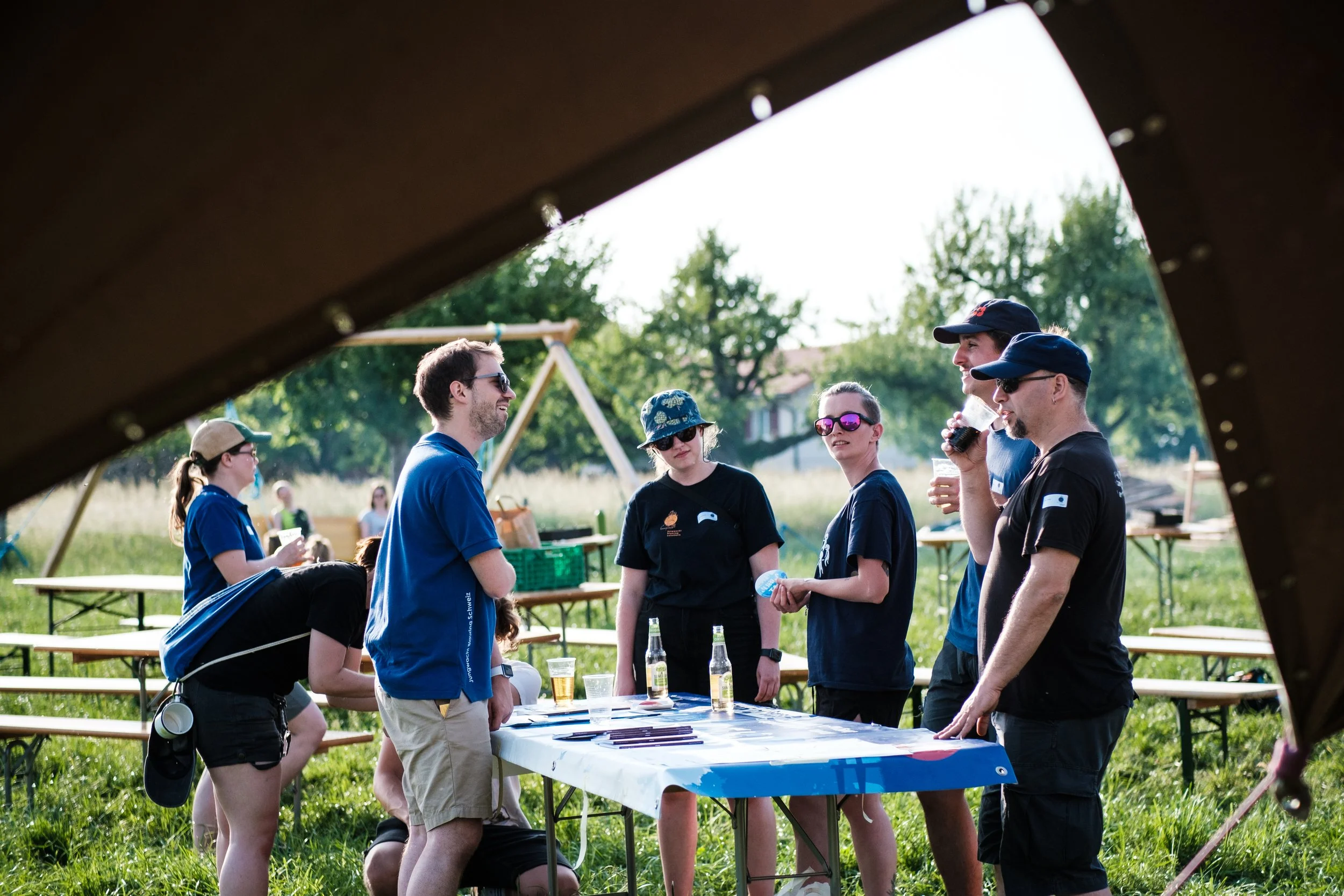 Gruppe junger Menschen bei einem Outdoor-Event, die um einen Tisch mit Papieren und Getränken stehen, im Park auf einer Wiese, mit Bäumen im Hintergrund.
