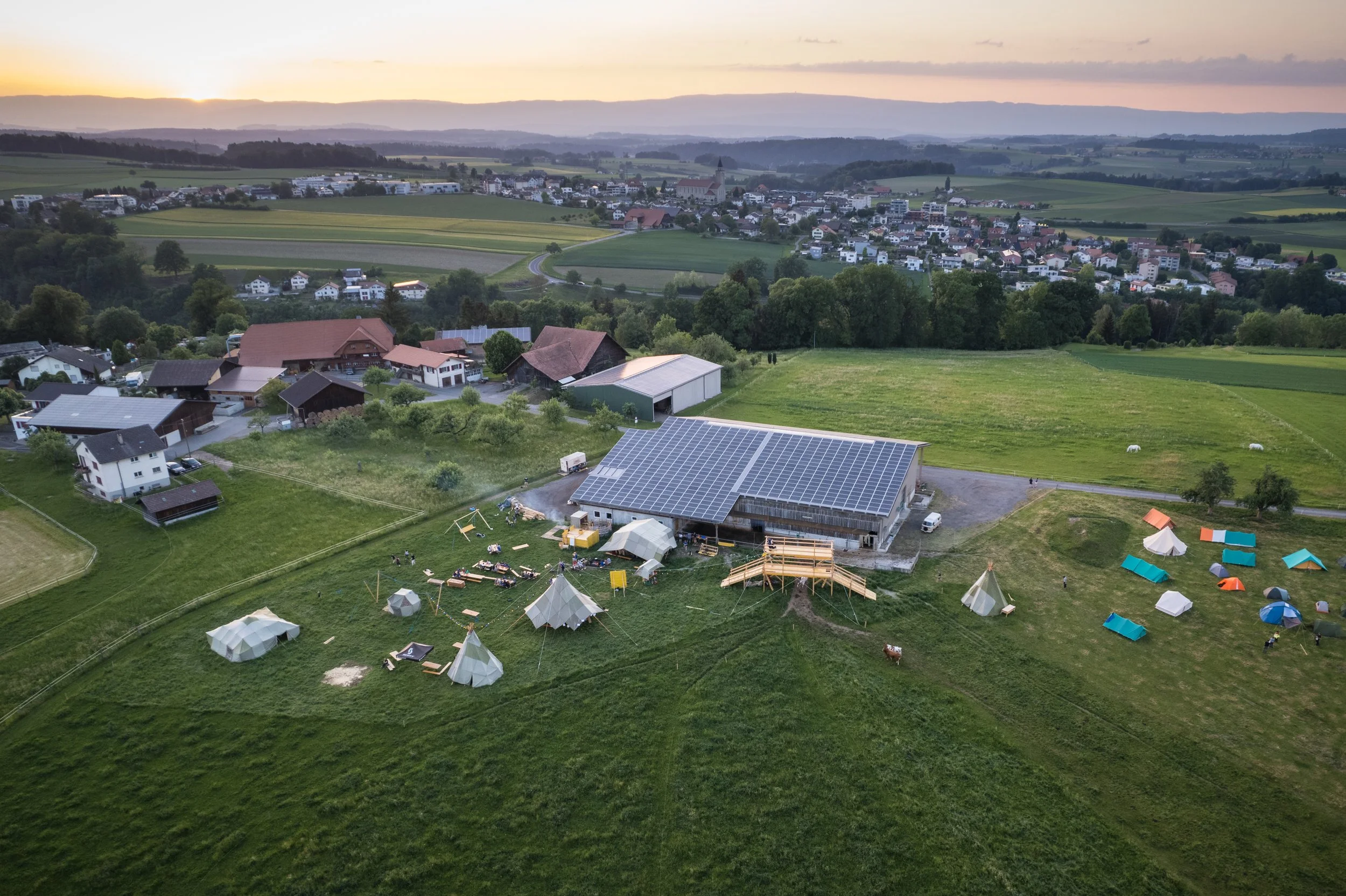 Luftaufnahme eines ländlichen Gebiets mit einem Gebäude, das Solarpanels hat, und Zelten im Freien bei Sonnenuntergang.
