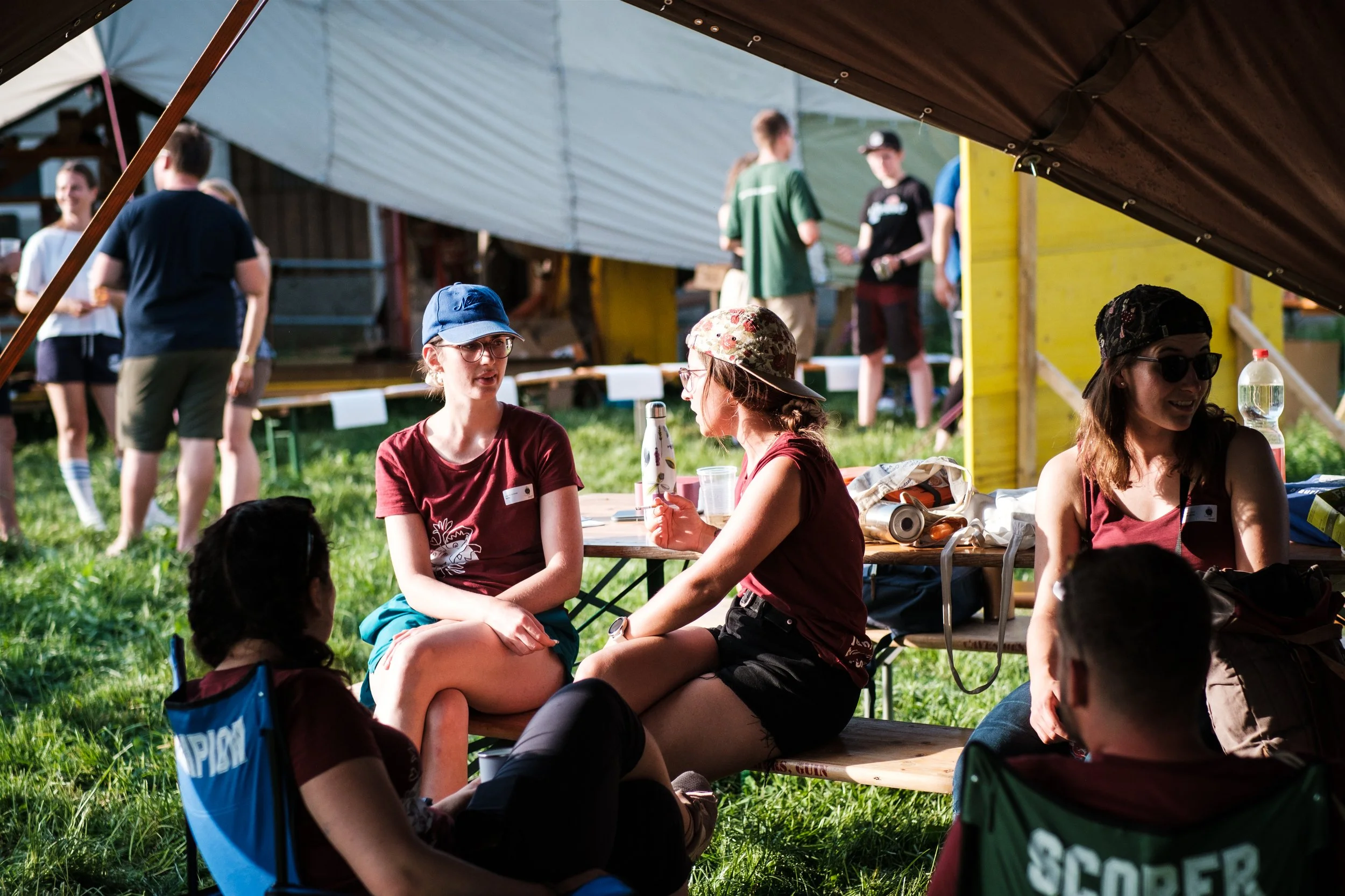 Jugendliche im Zeltlager, sitzen und unterhalten sich, Kinder im Hintergrund beim Spielen.