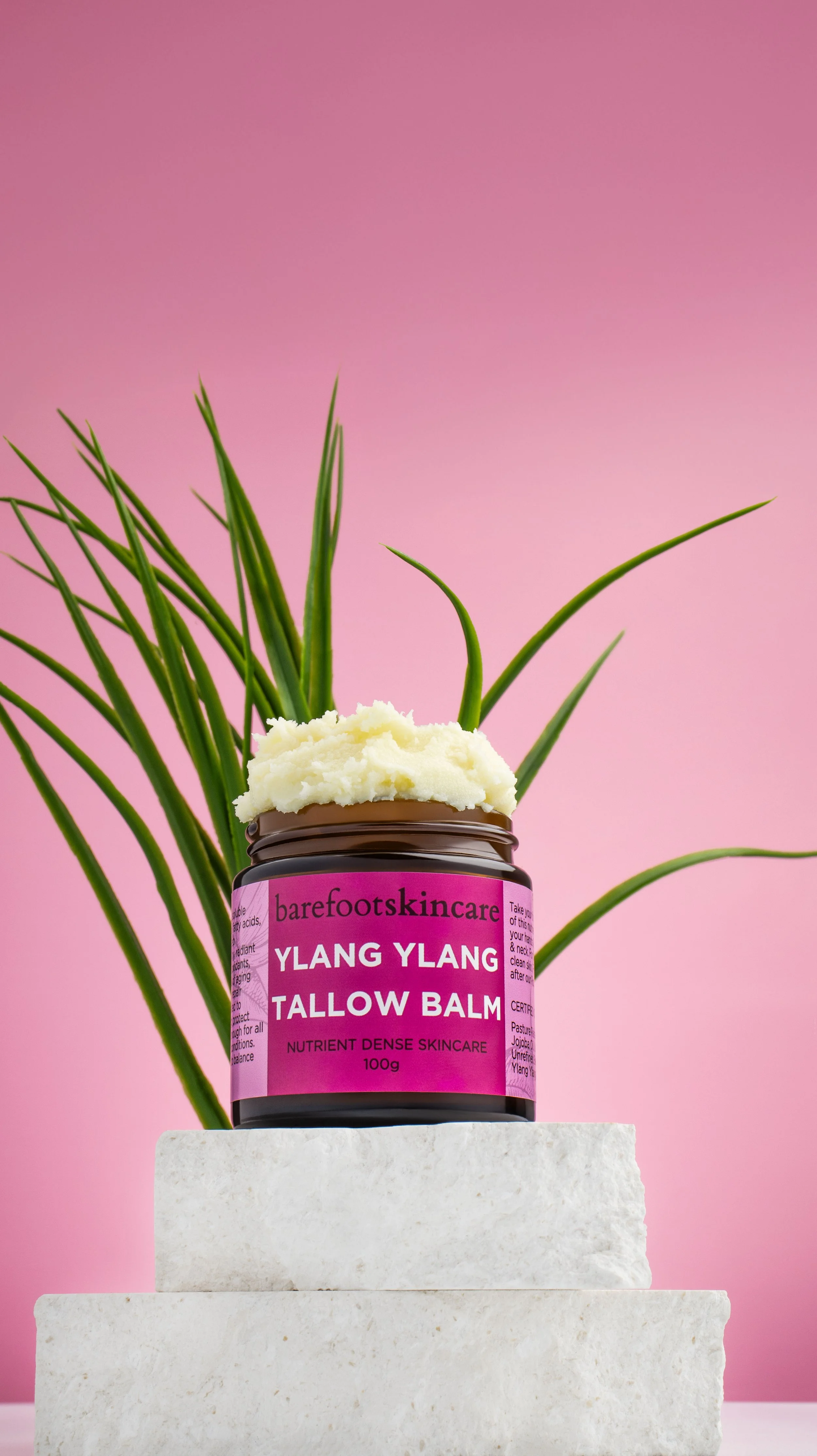 A jar of barefoot skincare Ylang Ylang Tallow Balm with scooped out product on top, placed on white stacked blocks with a green plant in the background against a pink backdrop.