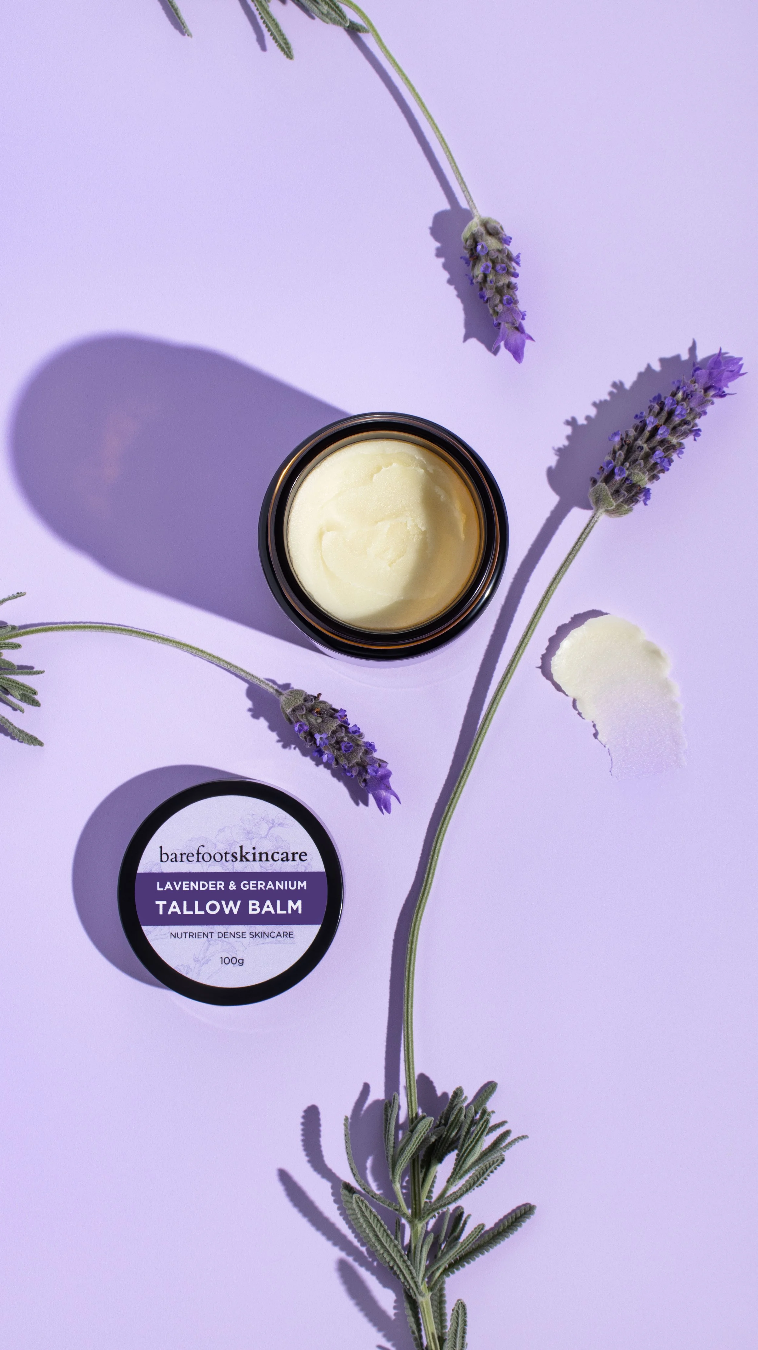 A jar of barefoot skincare lavender and geranium tallow balm, with sprigs of lavender and geranium flowers on a light purple background.