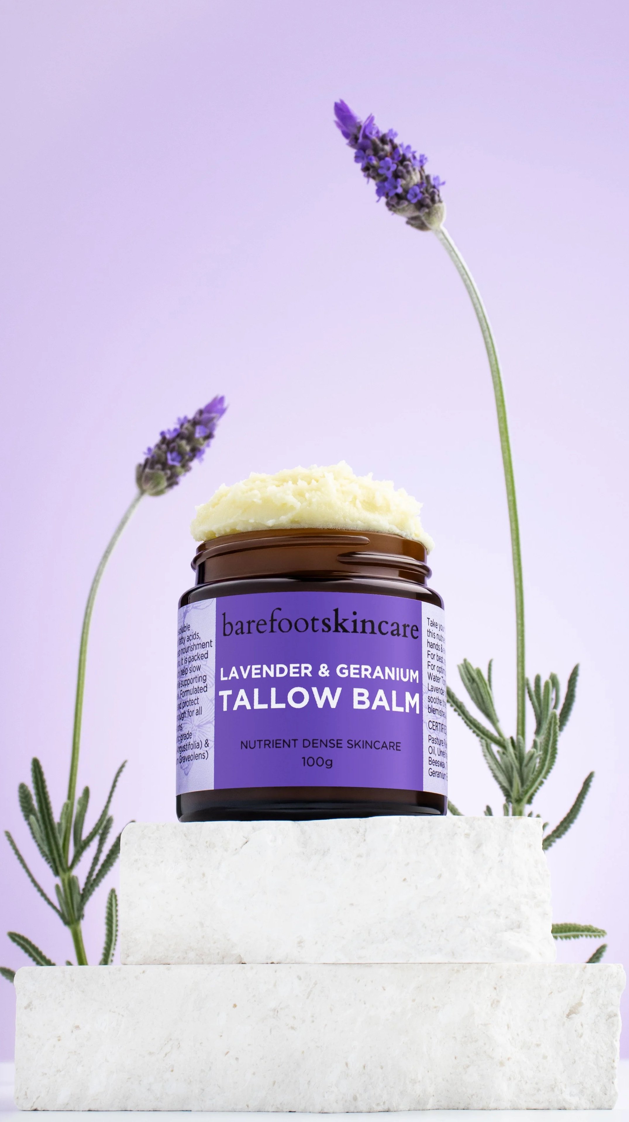 A jar of Barefoot Skincare Lavender & Geranium Tallow Balm placed on a white stone block, with lavender flowers and green leaves in the background, against a light purple backdrop.