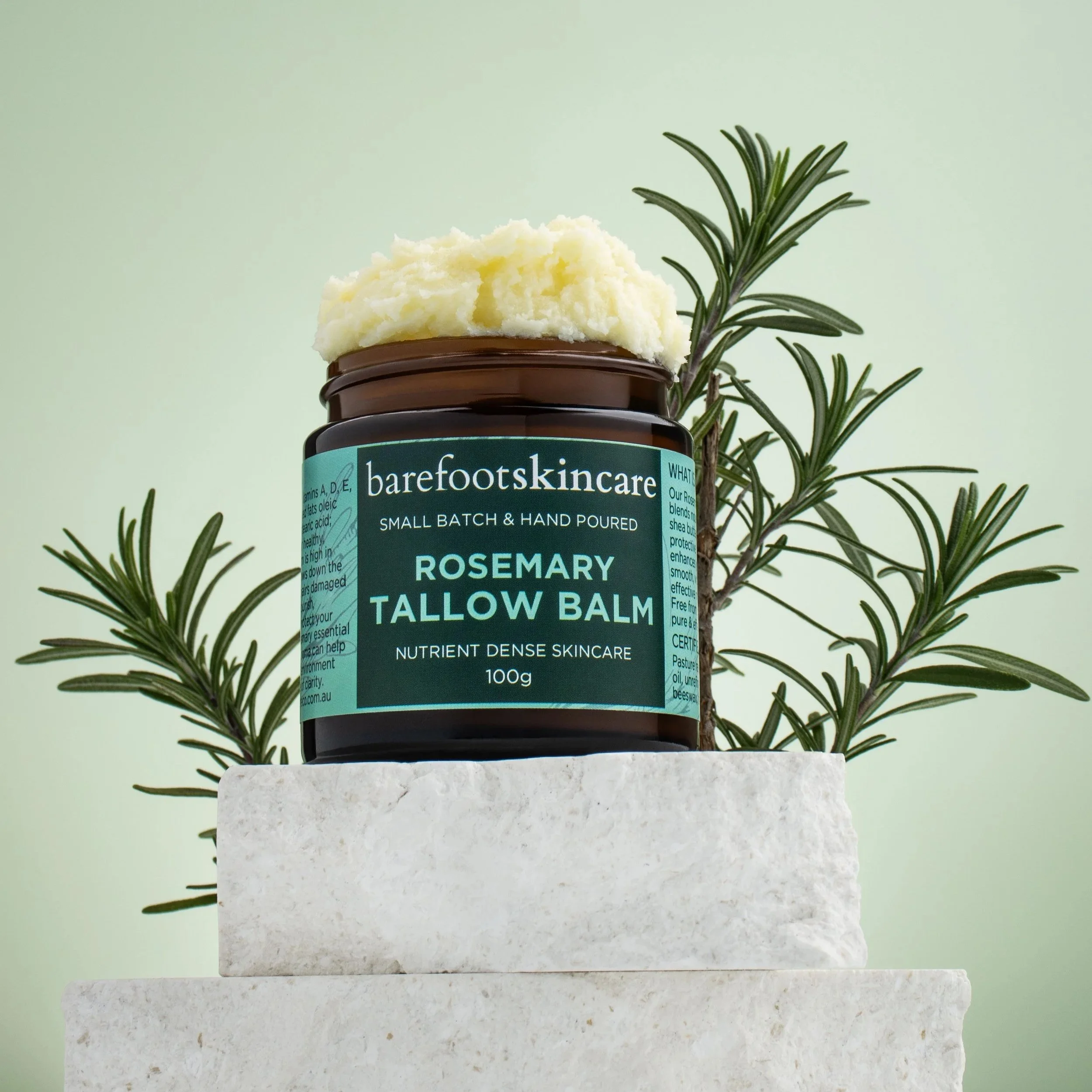 A jar of Barefoot Skincare Rosemary Tallow Balm with a dollop of balm on top, placed on white stone blocks with a sprig of rosemary behind it, set against a light green background.