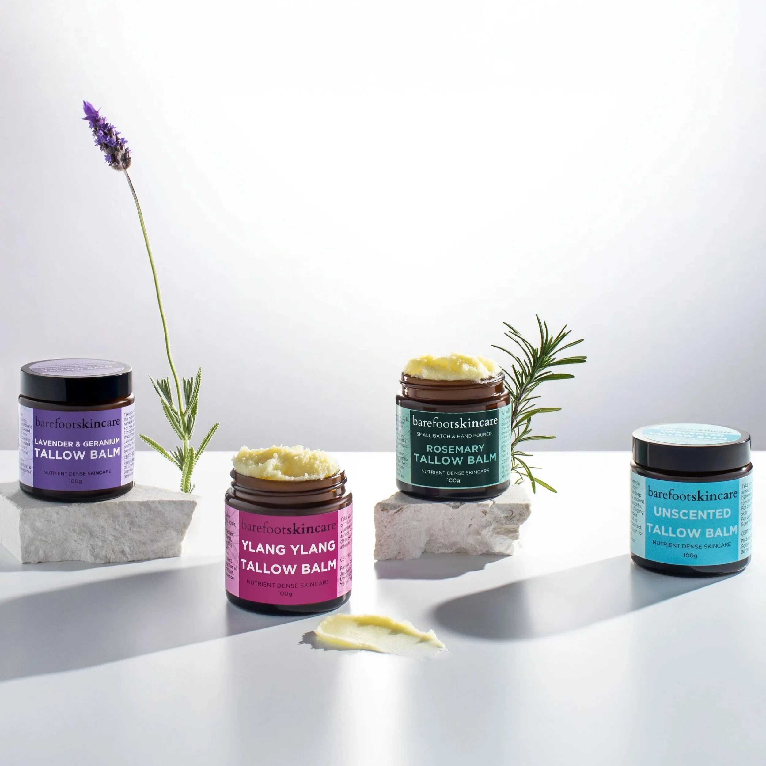 Five colorful jars of skincare with labels, some with open balm, set on stone slabs with sprigs of lavender and rosemary, against a white background.