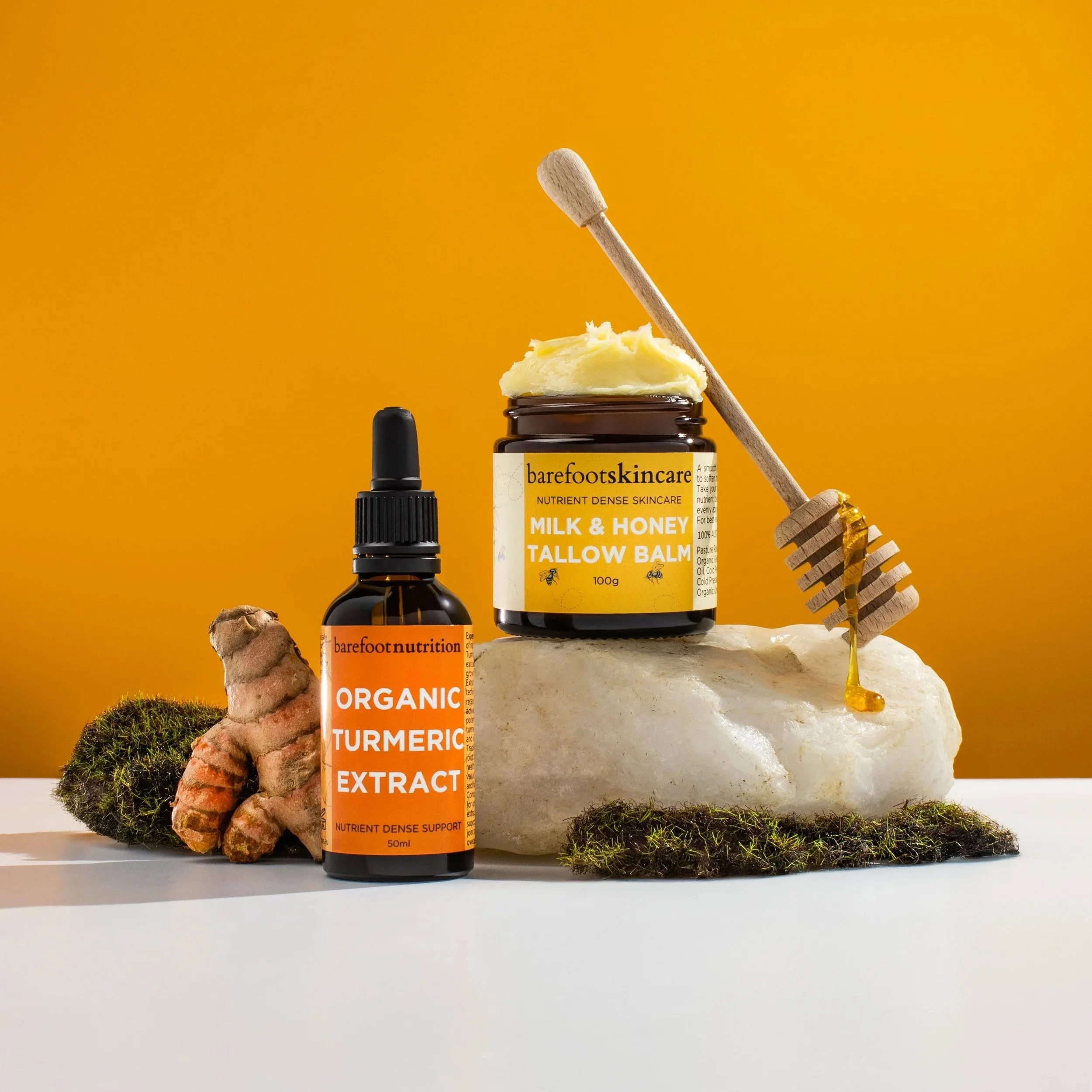 Jar of mango butter, container of milk and honey tallow balm, and bottle of organic turmeric extract with turmeric root and moss on a white surface against a yellow background.