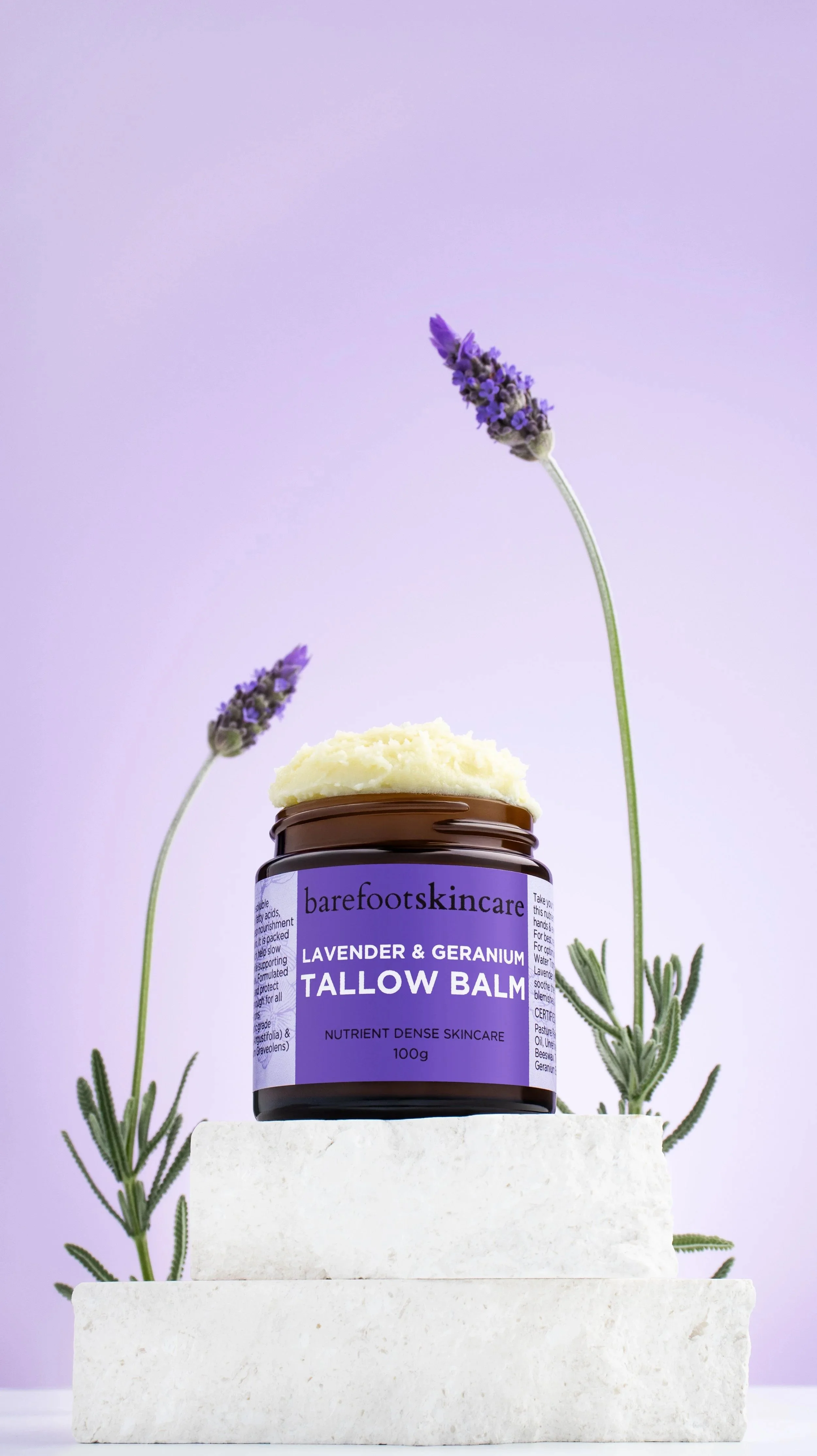 A small jar of lavender and geranium Tallow Balm from Barefoot Skincare placed on white stone blocks with lavender sprigs behind it and a light purple background.