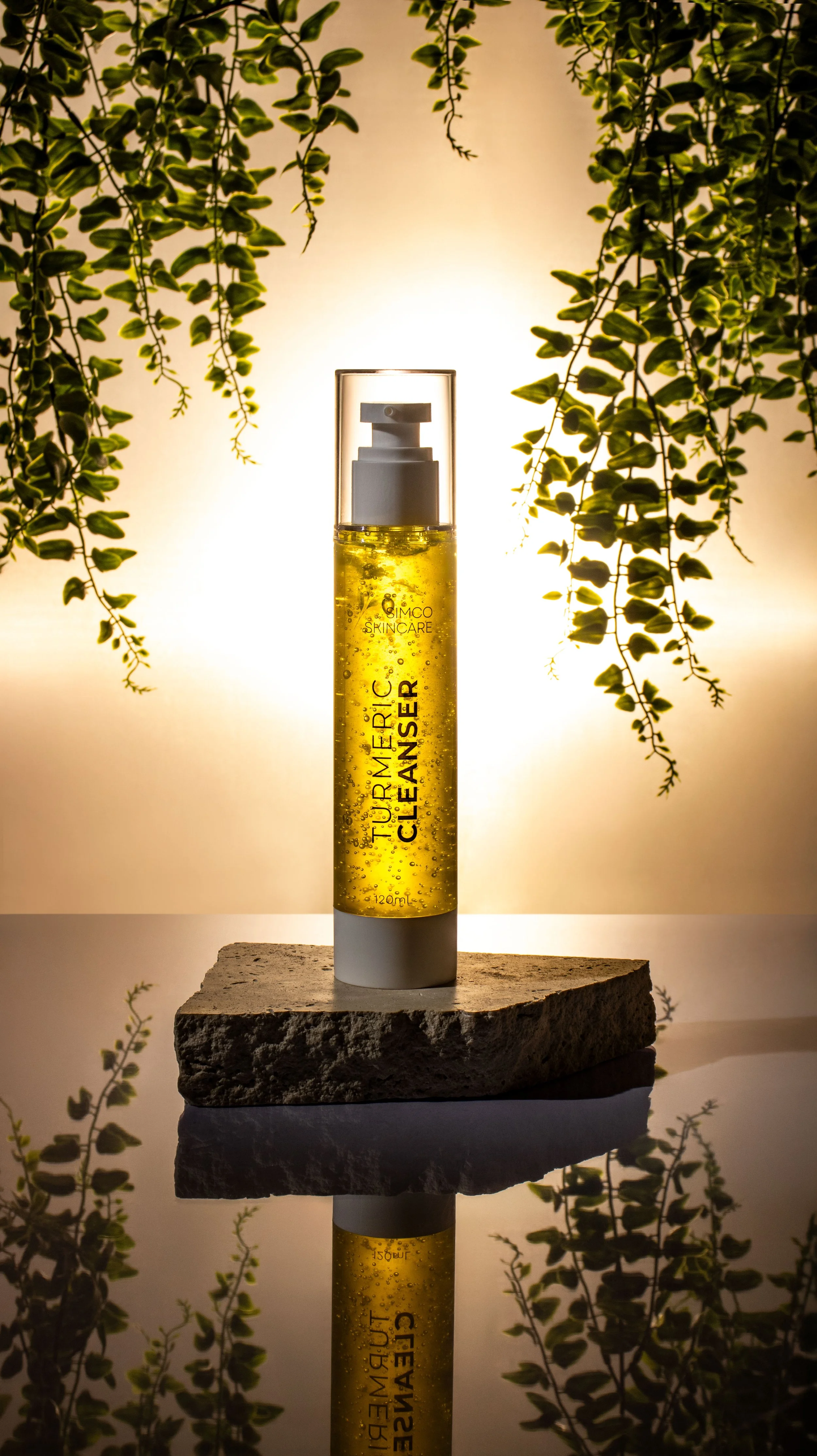 A bottle of turmeric cleanser with a foaming pump sits on a stone slab, reflected in a mirror. The background is lit, with leafy green plants hanging around.