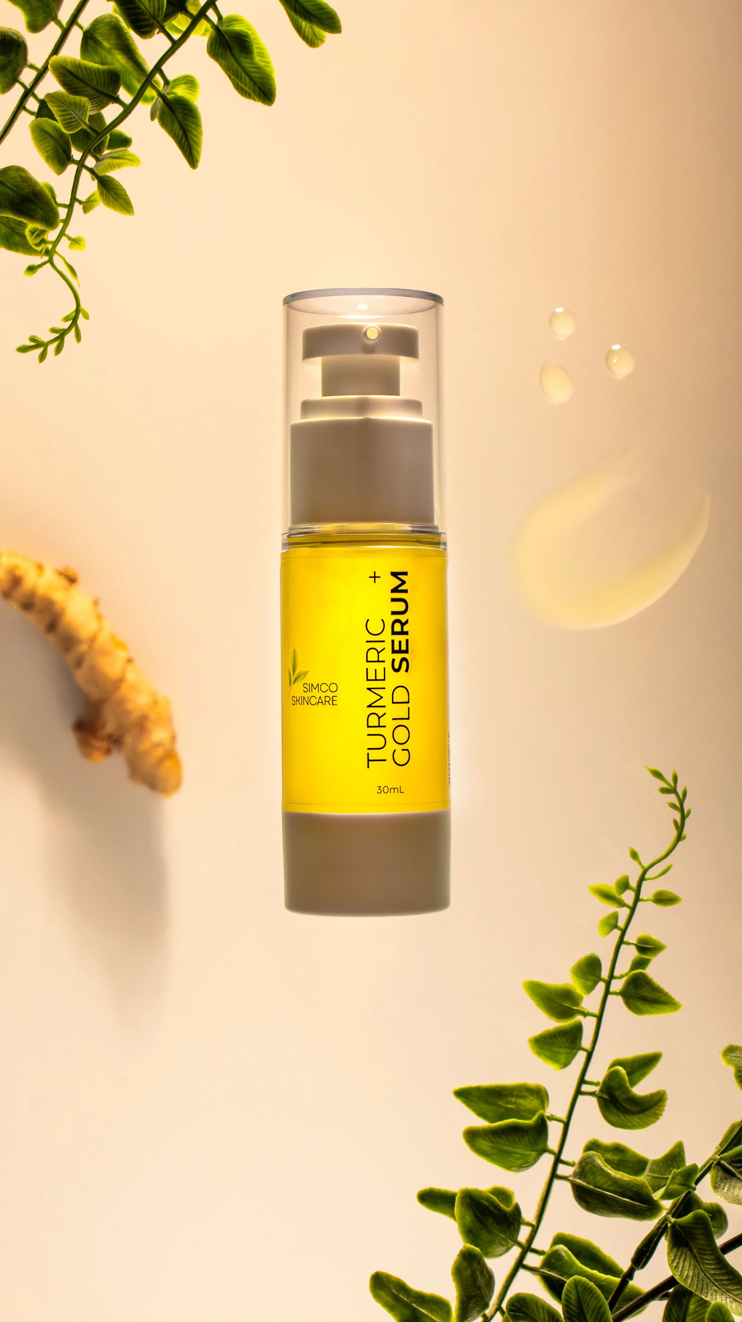 A bottle of turmeric and gold serum with a white pump dispenser, surrounded by green plant leaves, on a light background.