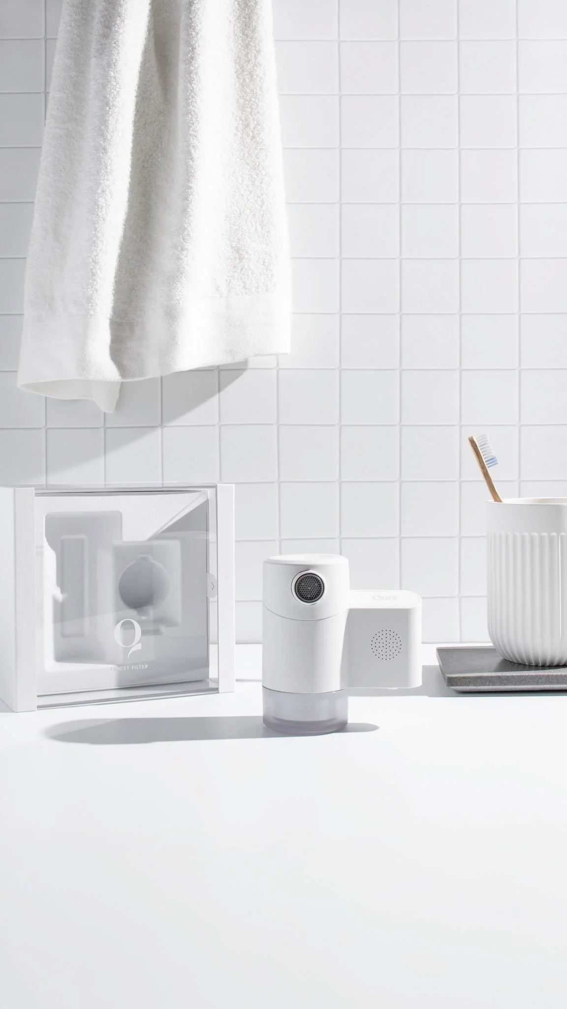 A white countertop with a small spray water diffuser, a box containing a water filter, a white toothbrush with blue bristles in a white, textured cup, and a gray tray. A white towel hangs on a tiled wall in the background.