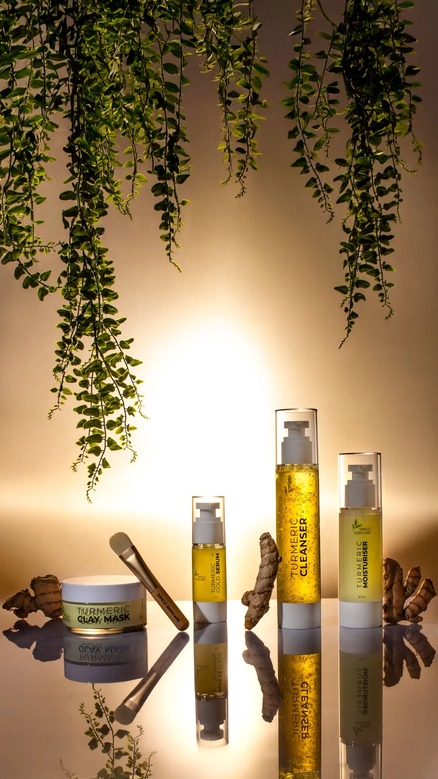 Collection of skincare products including turmeric clay mask, old serum, toner, and moisturizer bottles arranged on a reflective surface with ginger roots and hanging greenery in the background.