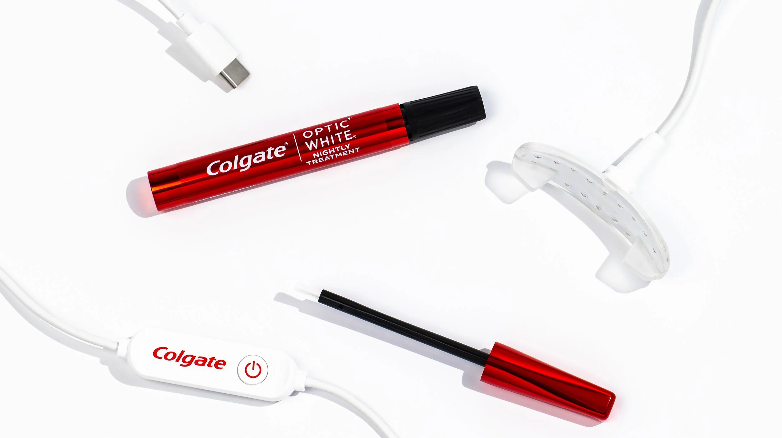 A white background with a red Colgate Optic White toothbrush marker, white earbuds, a black and red pen, and a white electronic device with a power button.