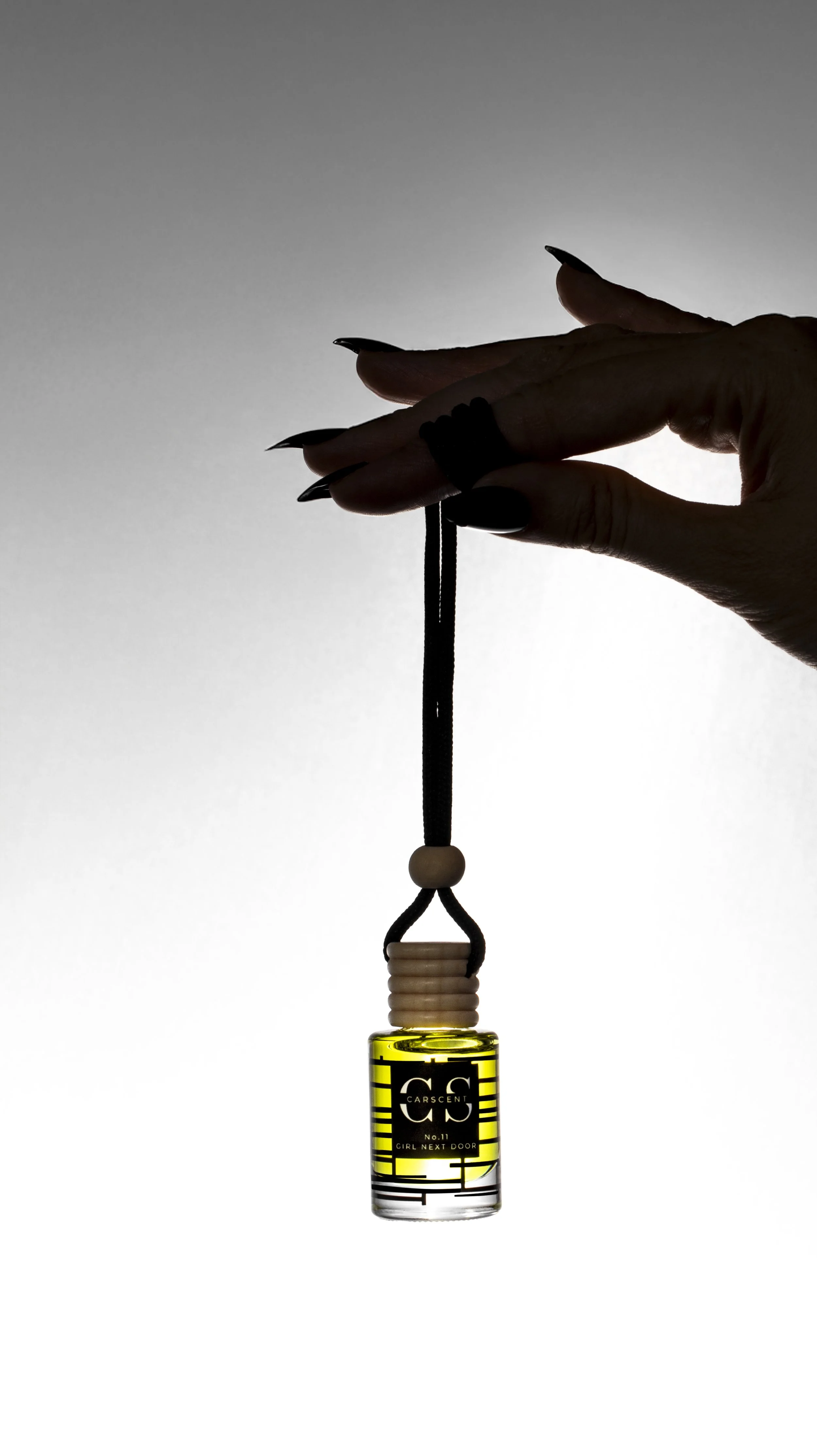 A hand with long black nails holding a small yellow perfume bottle with a black strap. The background is a gradient from light gray to white.