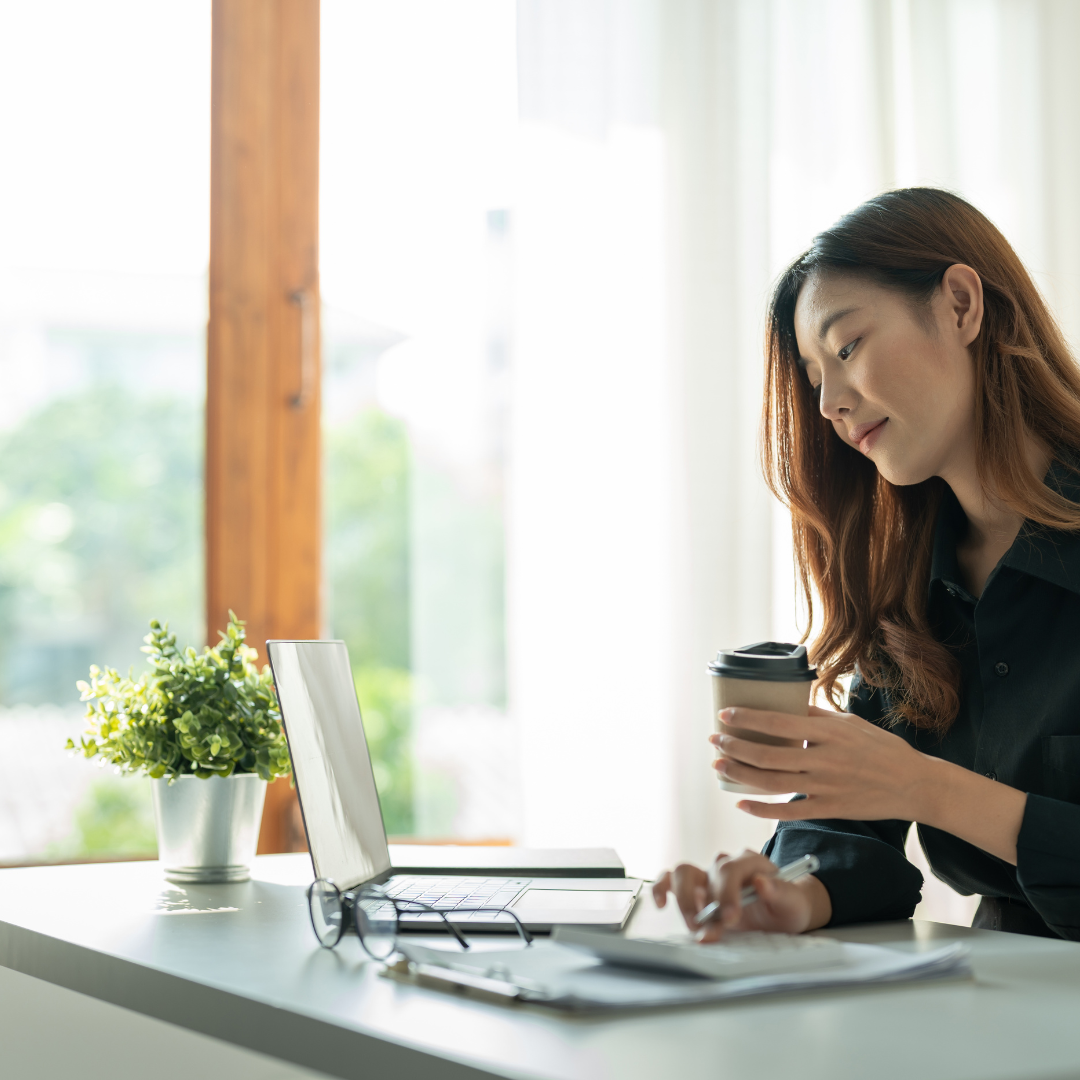 Woman sitting at a desk with a laptop, holding a coffee cup, in a bright room with a window and green plants.