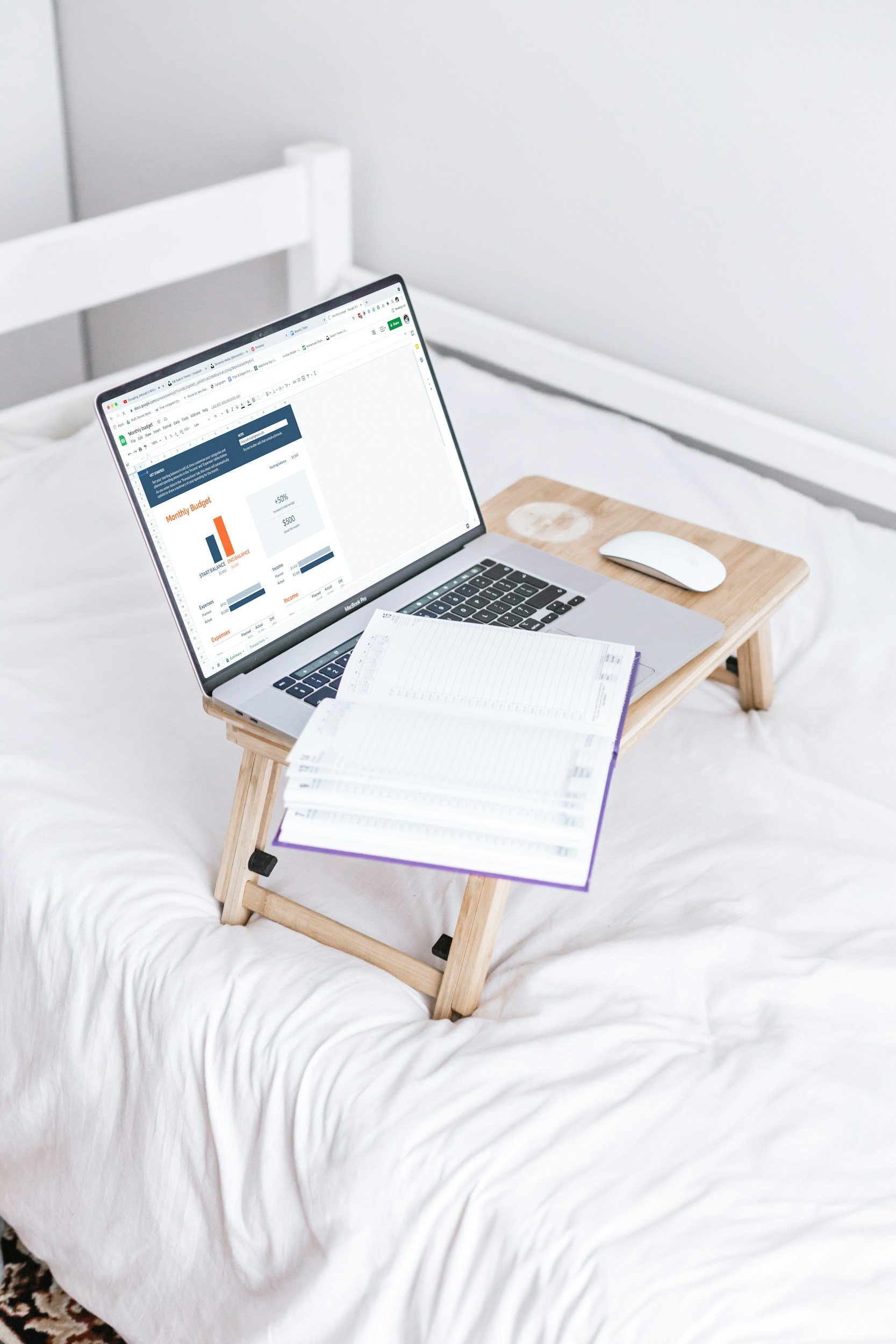 A laptop displaying a financial or budgeting spreadsheet on a wooden bedside table. There is a white computer mouse and an open notebook with handwritten notes on the same table, which is atop a white bed with a quilted bedspread.