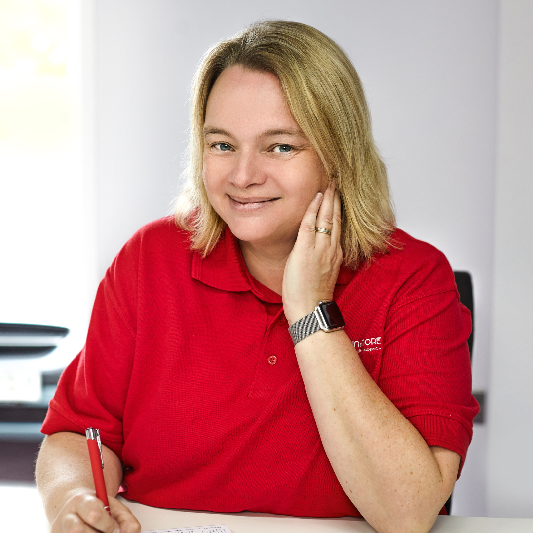 A woman with blonde hair wearing a red polo shirt smiling while sitting at a desk, holding a pen, with her left hand touching her face and wearing a smartwatch.