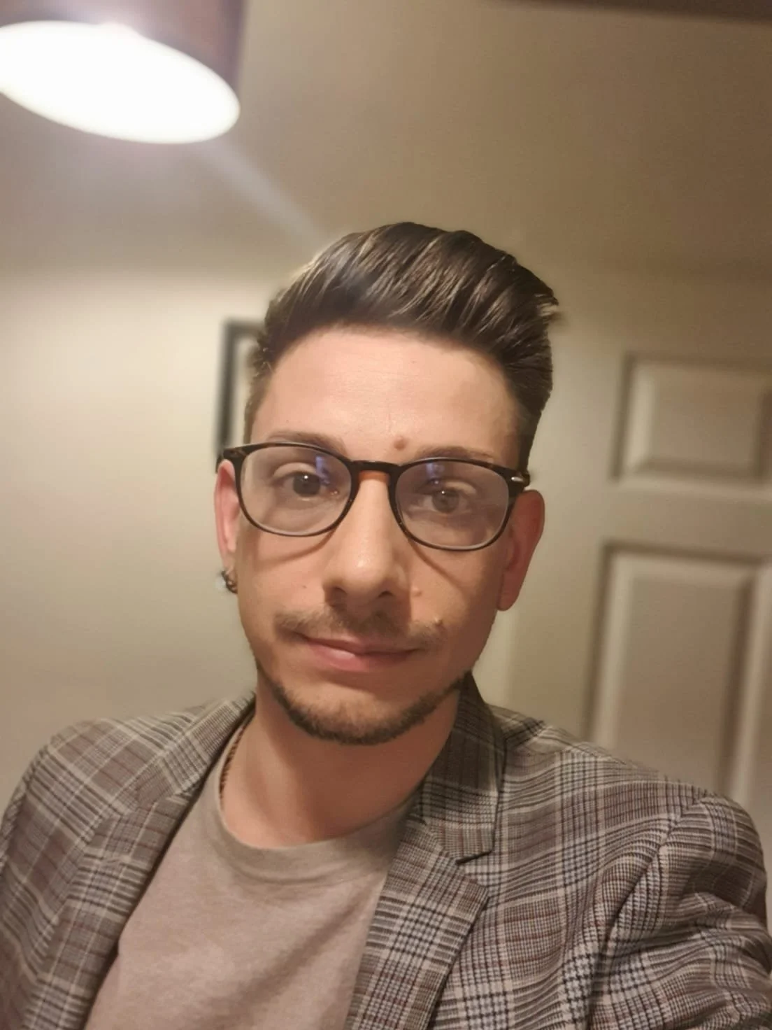 A young man with dark hair, glasses, and a light beard, dressed in a plaid blazer and light shirt, taking a selfie indoors, with a blurred background and warm lighting.