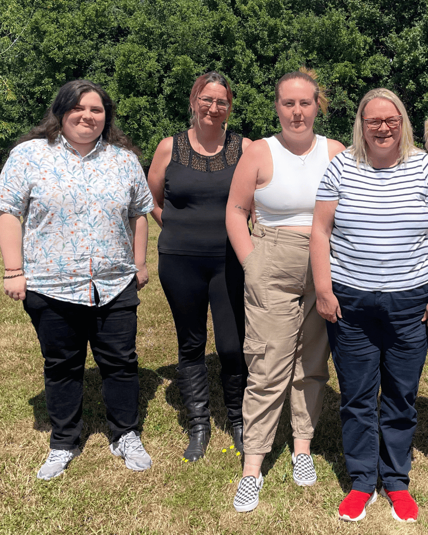 Four women standing outdoors on grass in front of green trees, smiling at the camera.