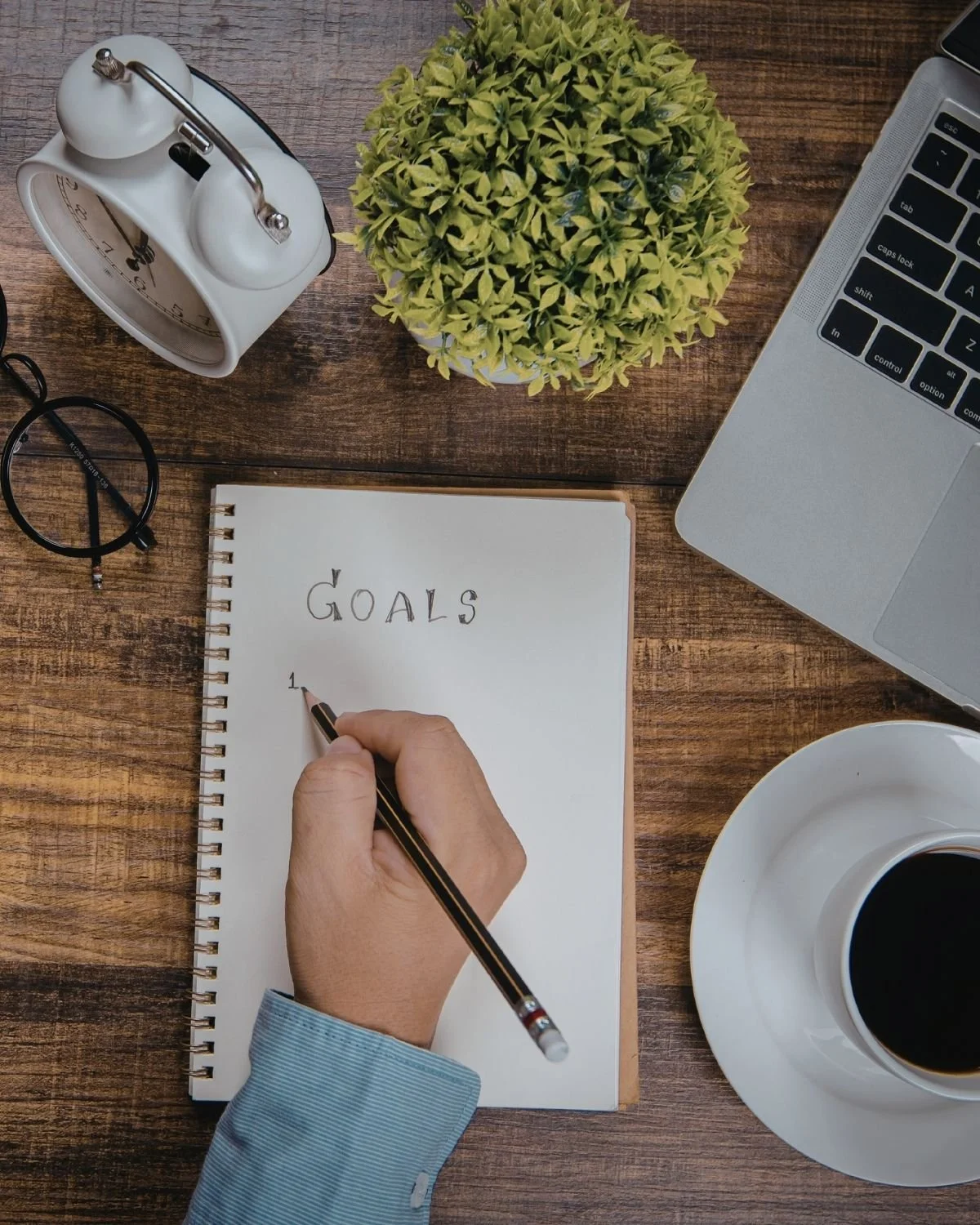 Top view of a wooden desk with a person's hand writing 'Goals' in a notebook, surrounded by a clock, a plant, a pair of glasses, a laptop, a cup of coffee, and a white mug.