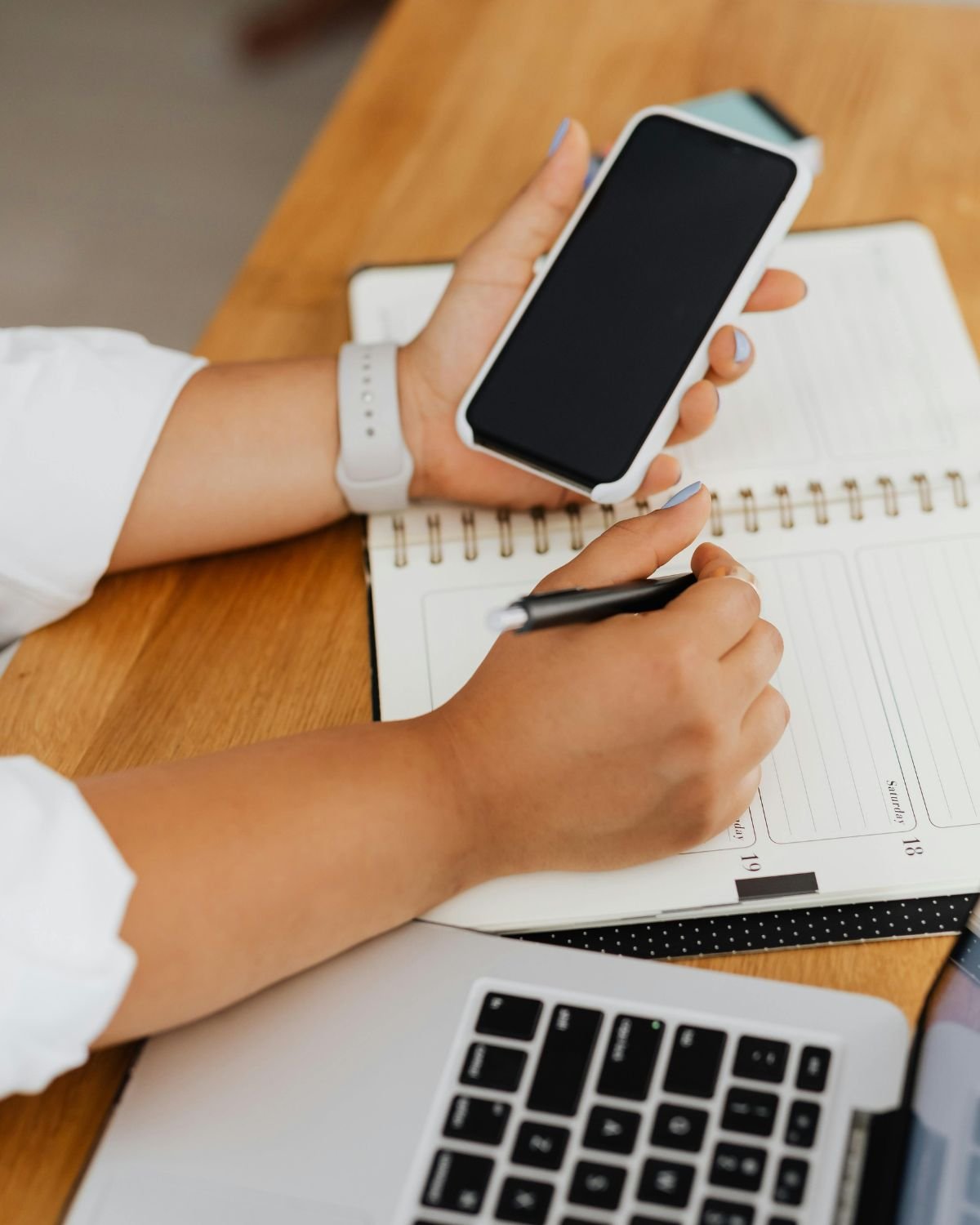 A person holding a smartphone in one hand and writing in a notebook with a pen in the other hand, on a wooden table with a laptop nearby.