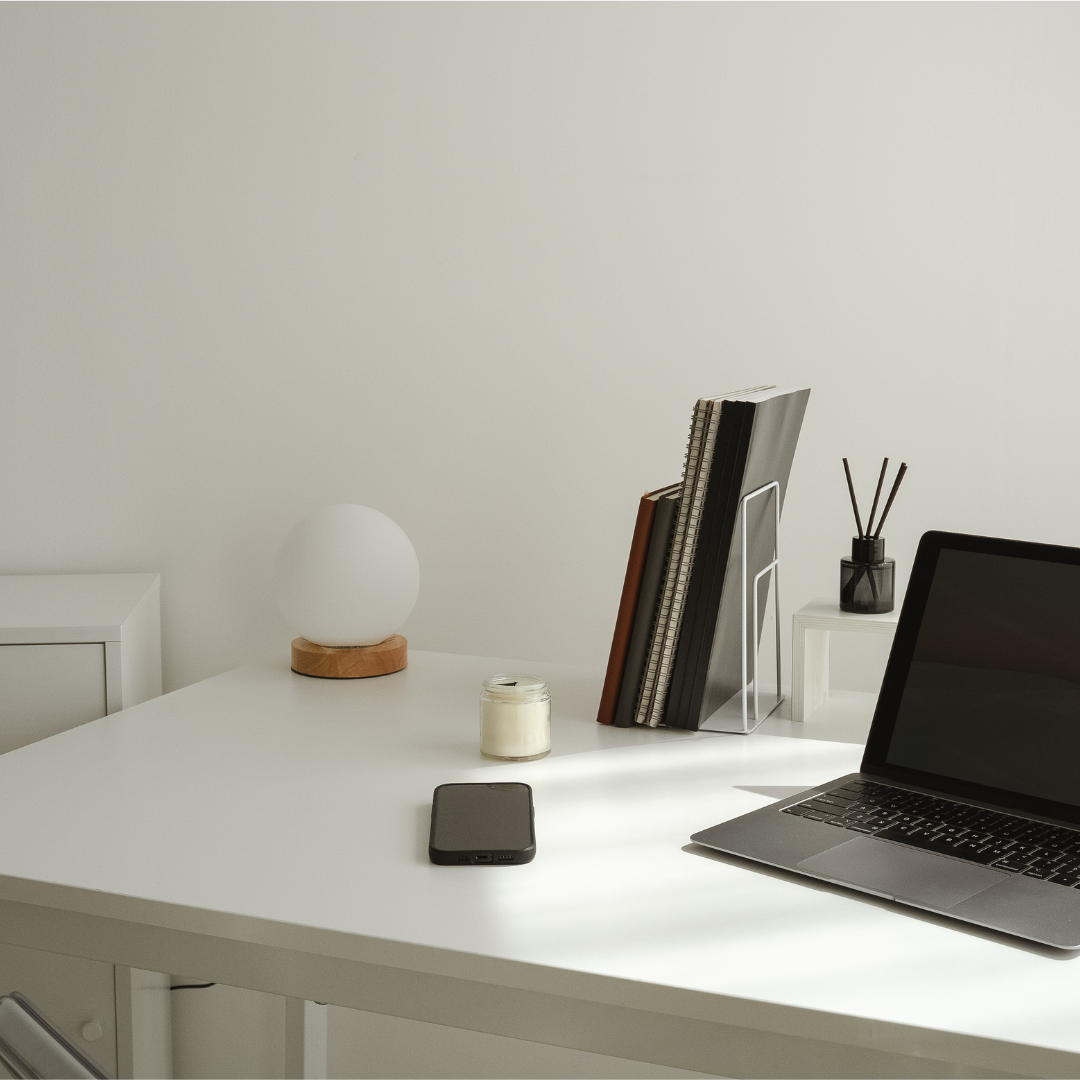 Minimalist white desk with a laptop, a smartphone, a candle, a modern lamp, a book holder with notebooks and papers, and a small vase with reed diffuser sticks.